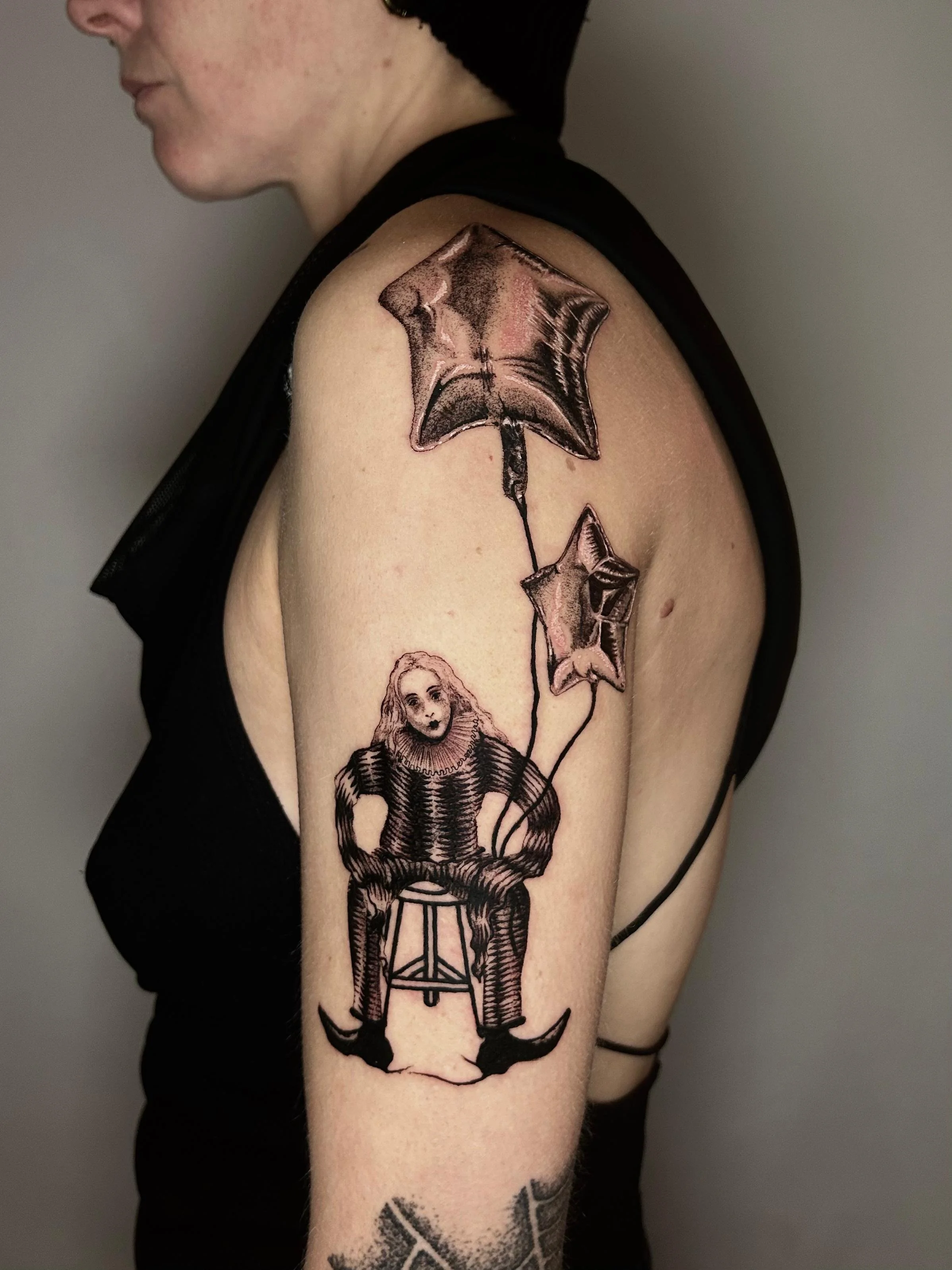 Tattoo auf dem Oberarm einer Frau: Ein Junge sitzt auf einem Stuhl, hält zwei Ballons, die in Form von Sternen gestaltet sind.