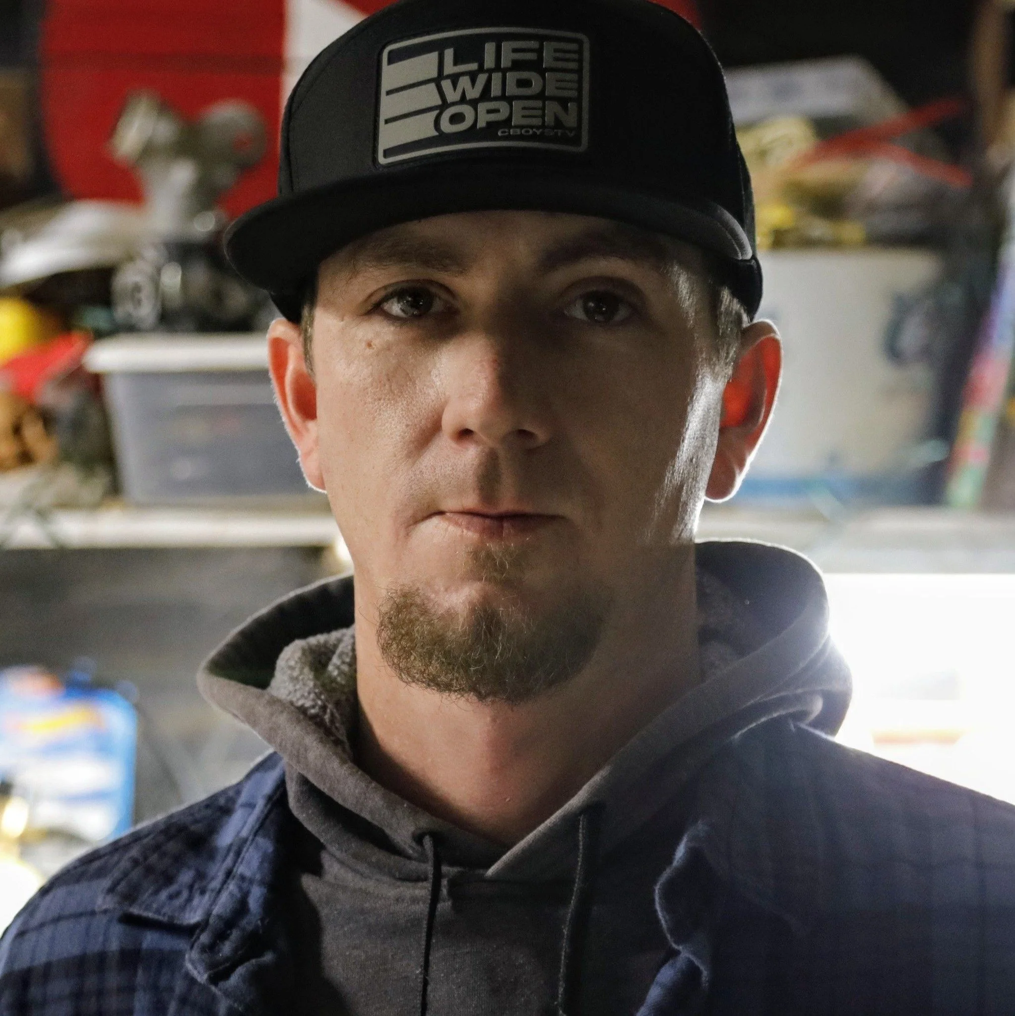 Portrait of a man wearing a black cap with a logo reading 'LIFE WIDE OPEN' and a dark hoodie, in a cluttered workshop or garage.