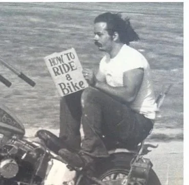 A man with long hair and a mustache sitting on a small motorcycle, holding a sign that reads 'How to Ride & Bike'.