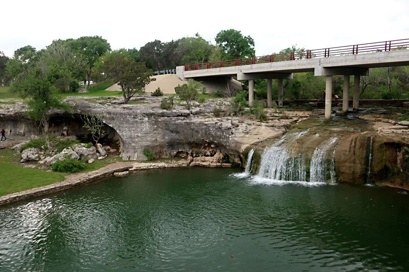 A small waterfall flowing into a green pond with surrounding rocks and trees, a bridge overhead, and a park area in the background.