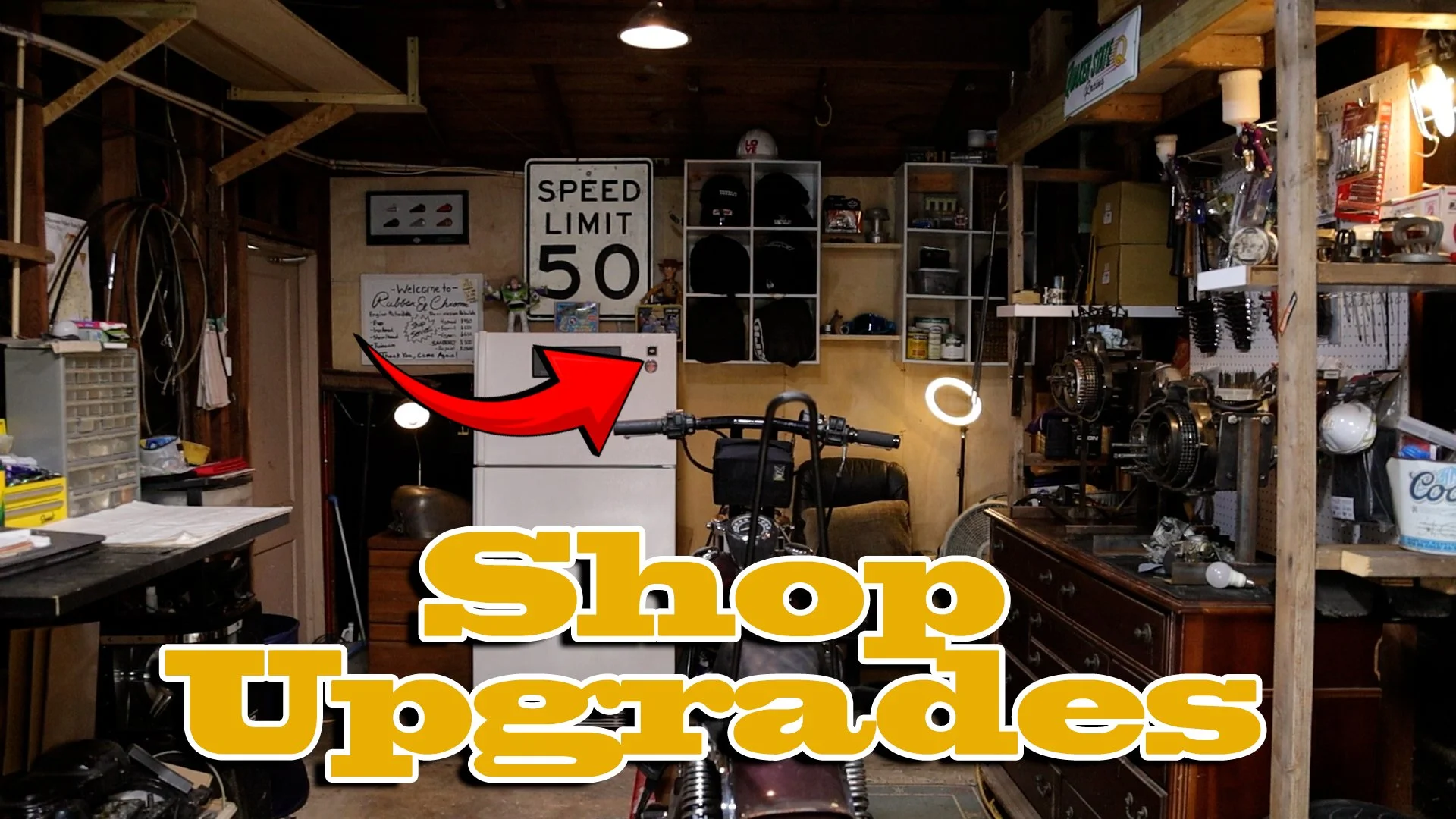 An interior view of a cluttered garage or workshop with various tools, shelves, and items. A white fridge with a small black box on top, a motorcycle, and a wooden cabinet are visible. There's a street sign that says 'Speed Limit 50,' a welcome sign, helmets, and other garage supplies. The text 'Shop Upgrades' is overlaid in bold yellow letters with a red arrow pointing towards the fridge.
