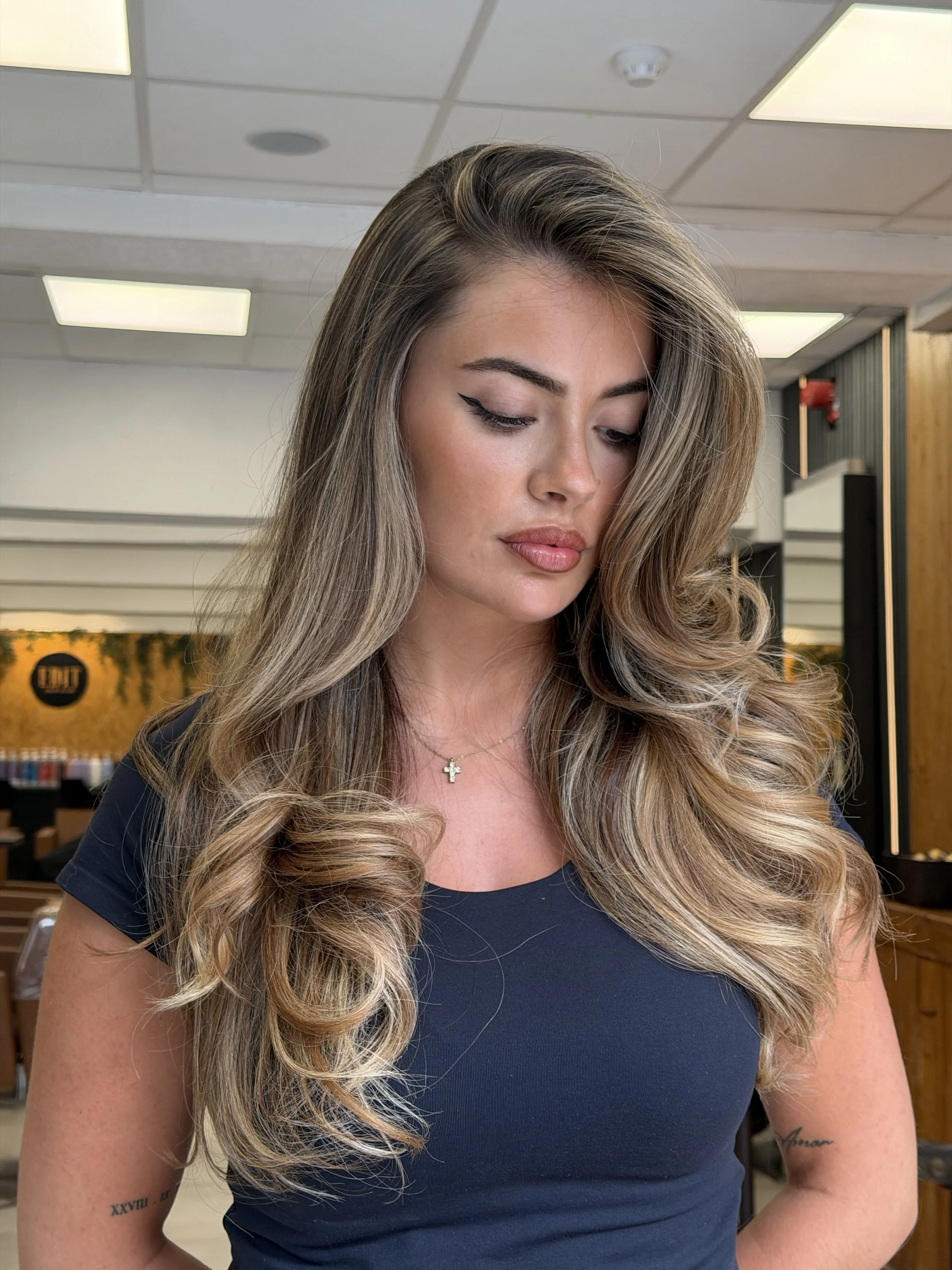 Close-up of a young woman with long, styled blonde hair, wearing makeup, a black top, a gold cross necklace, and tattoos on her arms, in an indoor setting with ceiling lights and wooden panels in the background.