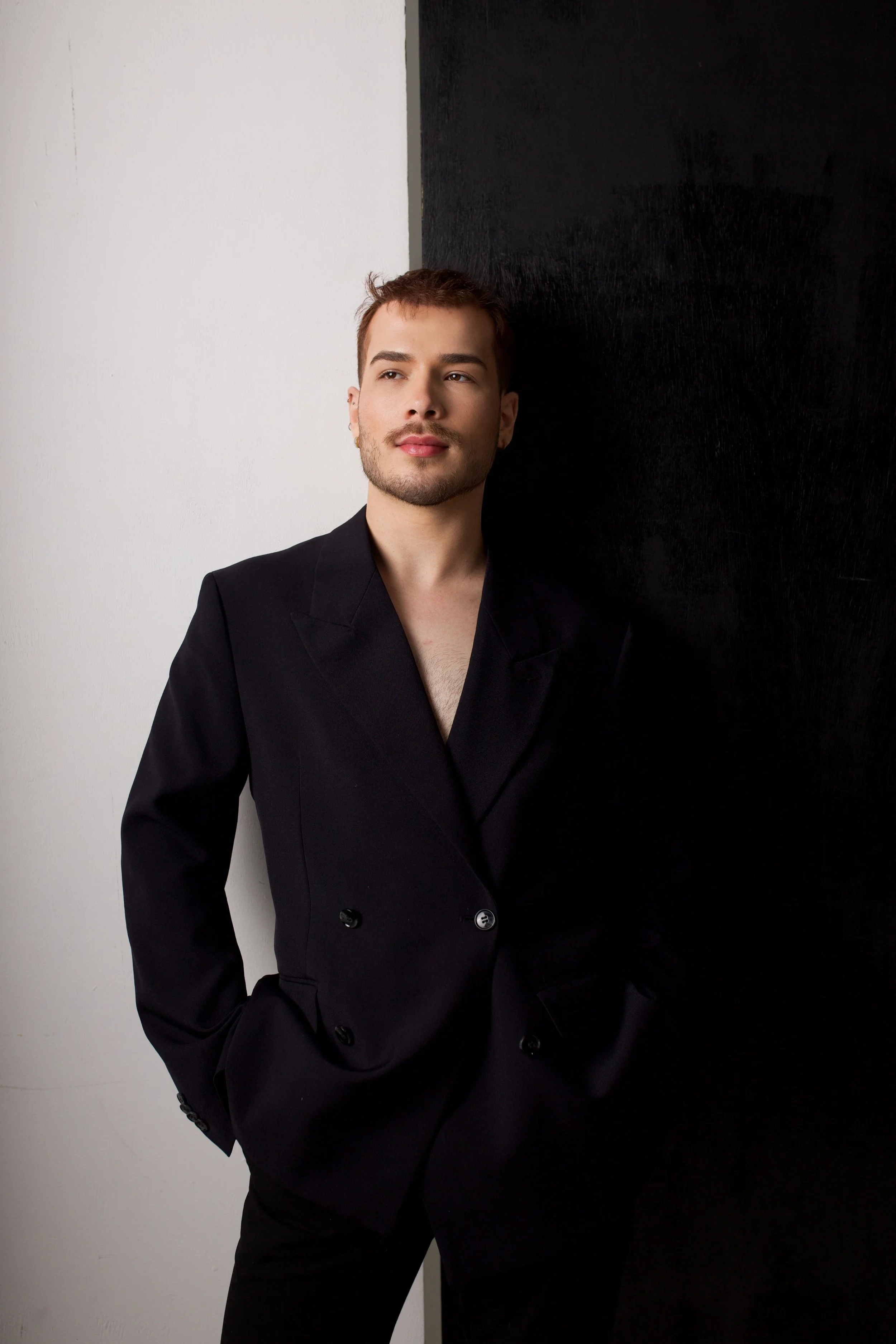 A young man with short brown hair and light facial hair, wearing a black blazer, stands against a background split into white and black sections, with a contemplative expression.