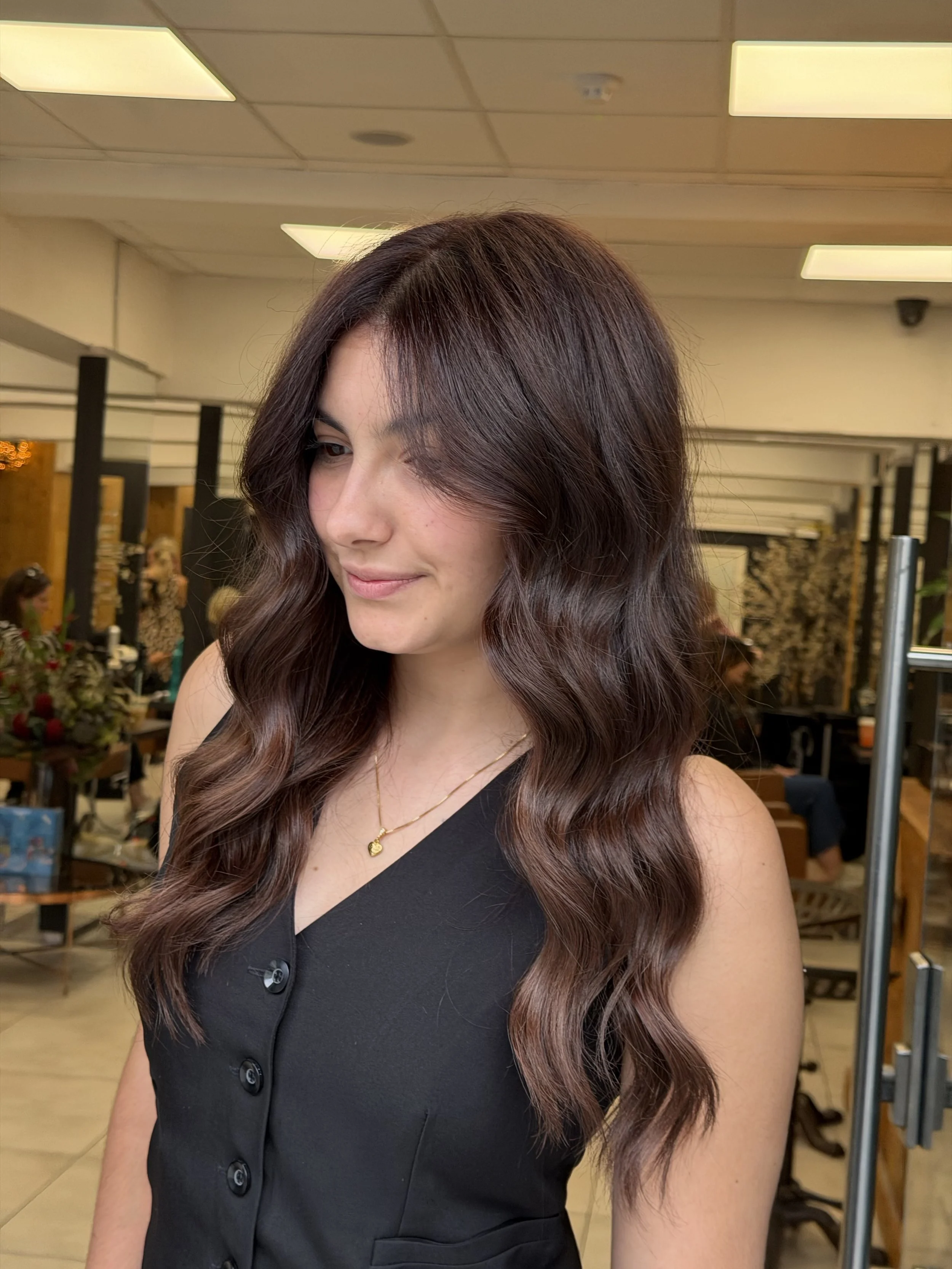 A woman with long, wavy brown hair and fair skin is standing in what appears to be a salon or waiting area. She is wearing a sleeveless black top with large buttons and a gold necklace with a heart-shaped pendant. The background features blurred people, floral arrangements, and salon equipment.