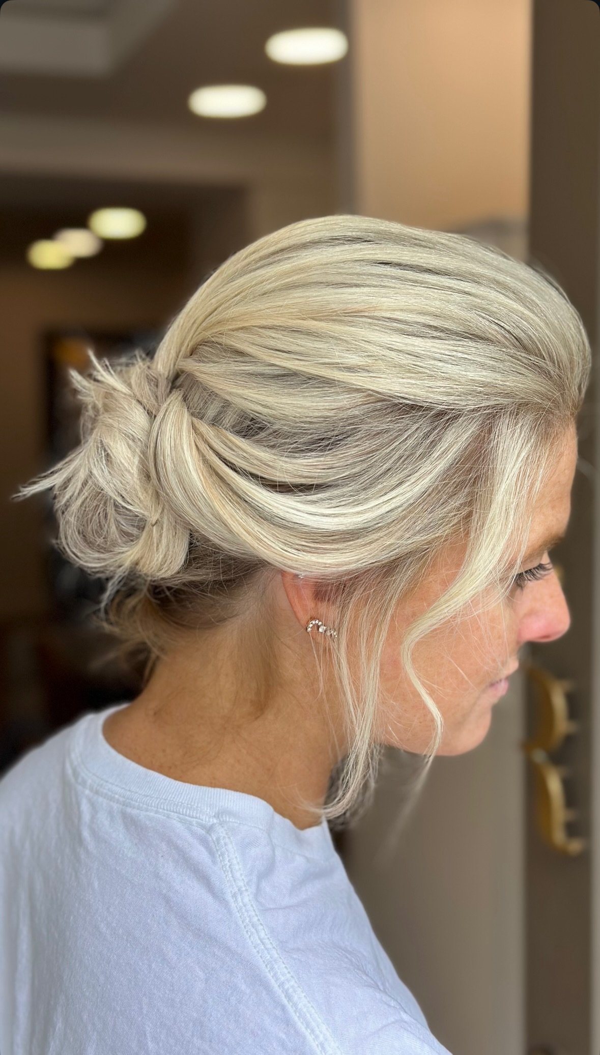 Side profile of a woman with platinum blonde hair styled in a low, twisted bun with loose curls, wearing a white top, and earrings.