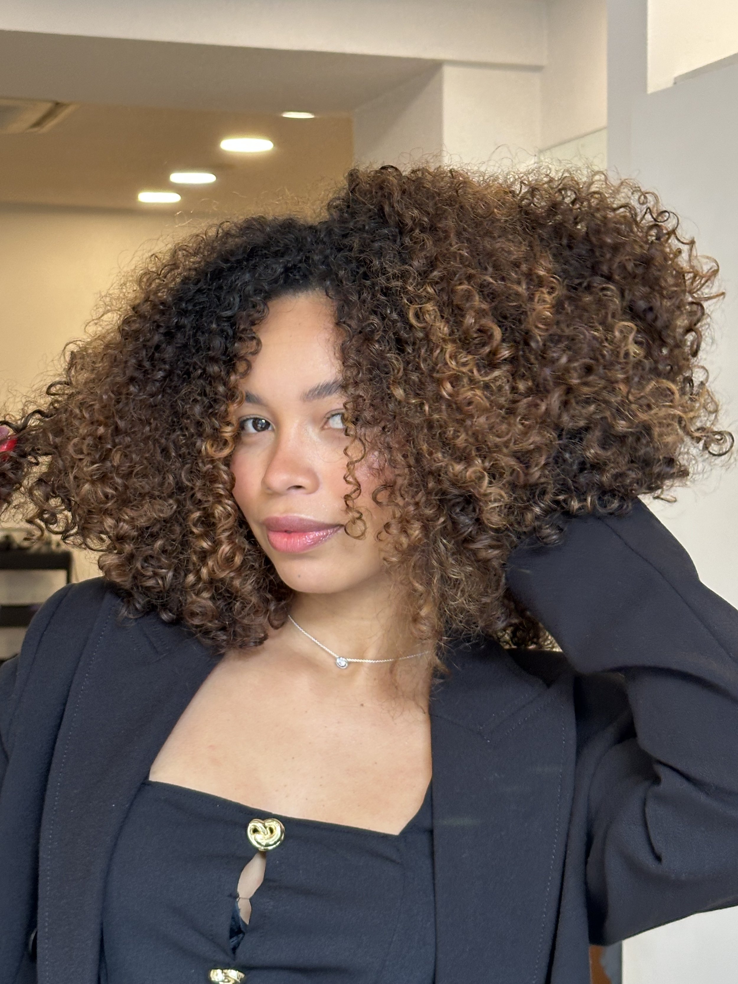 A woman with curly hair wearing a black blazer and a silver necklace, partially visible in the photo.