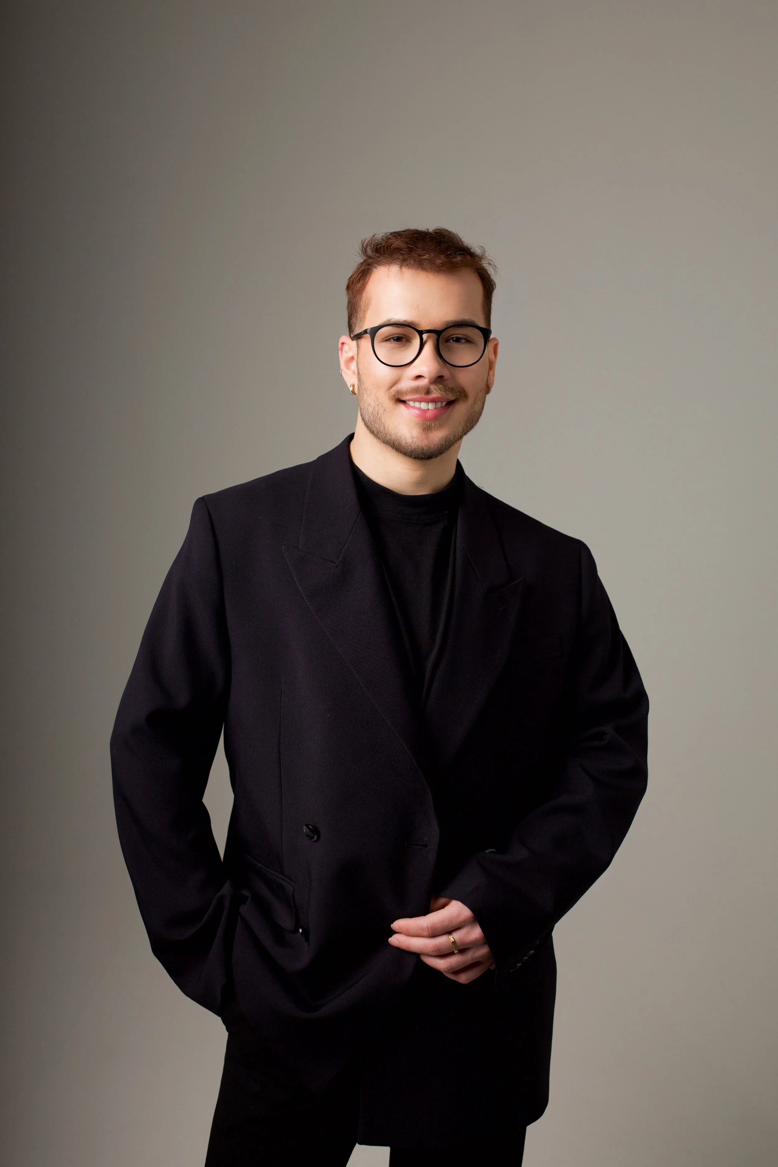 A young man with short brown hair, glasses, and earrings, wearing a black blazer and black shirt, standing against a plain gray background, smiling softly with his hands clasped in front of him.