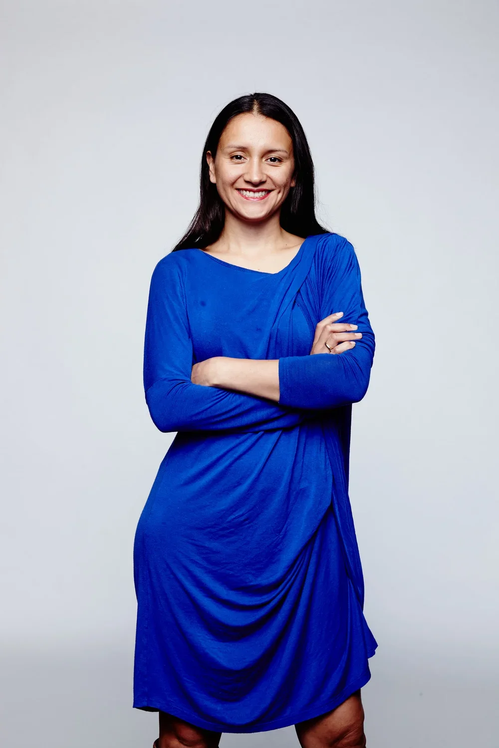 A woman with long dark hair wearing a bright blue dress, standing with her arms crossed and smiling against a plain white background.