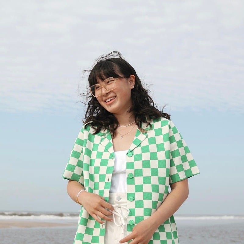 Young woman with glasses smiling on the beach, wearing a green and white checkered shirt over a white top.
