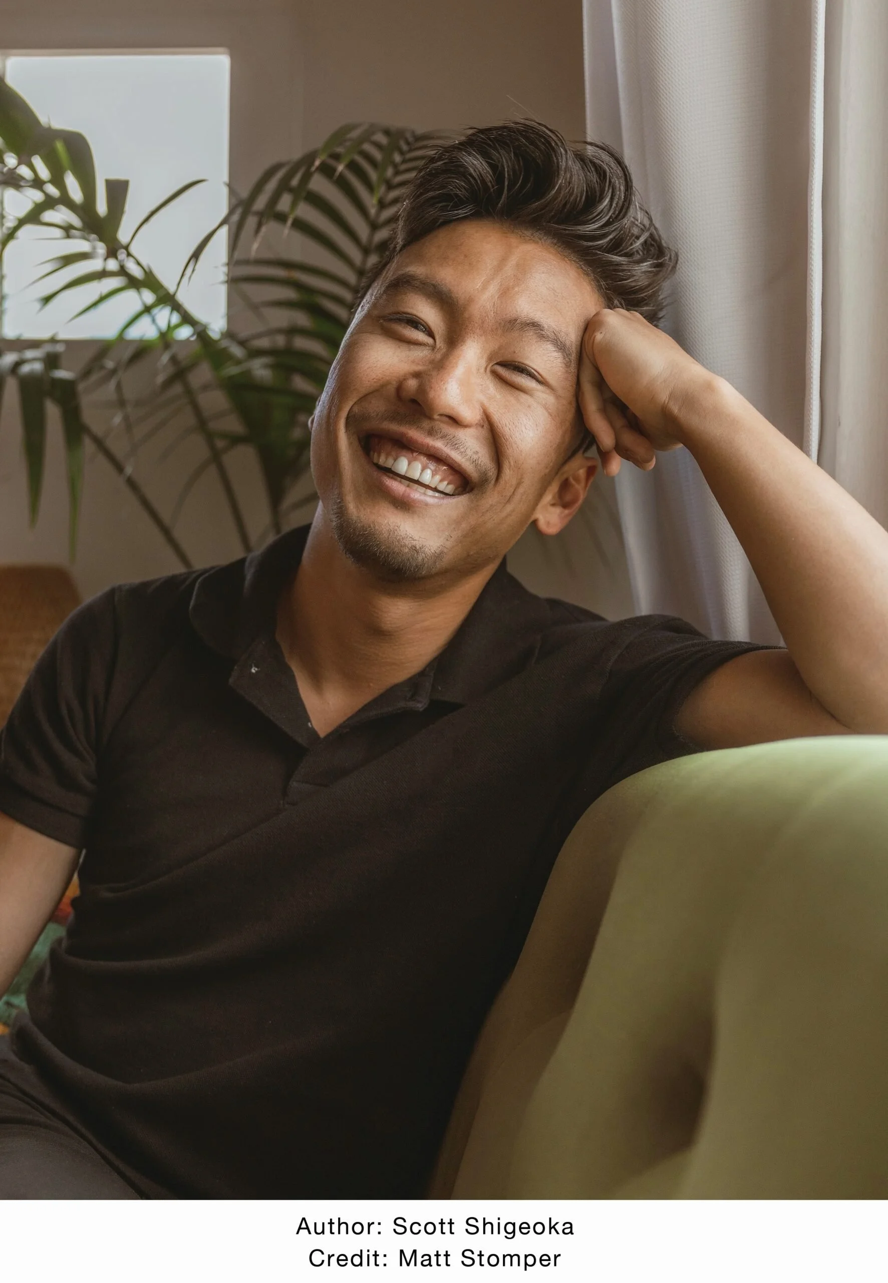 A smiling man sitting on a green couch, resting his head on his hand, with a large plant and a window with curtains in the background.