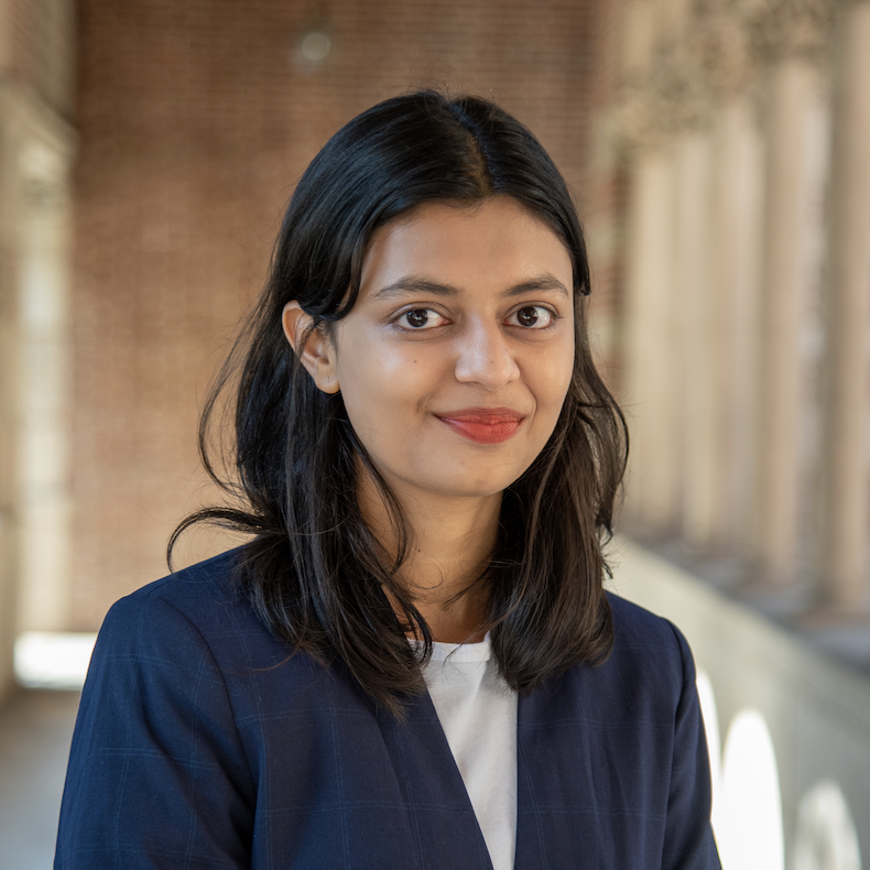 A woman with shoulder-length dark hair, wearing a navy blazer and white shirt, is standing outdoors with a brick wall and columns in the background.
