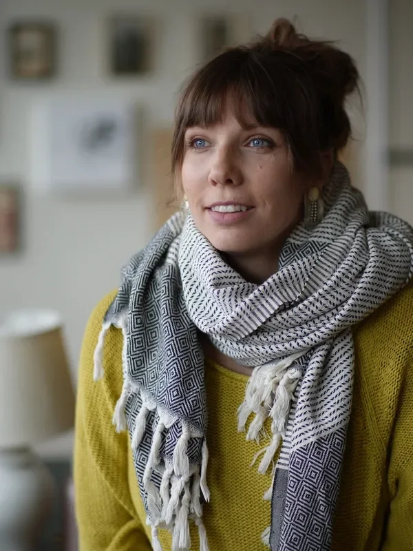 A woman with brown hair and blue eyes, wearing a yellow sweater and a patterned gray and white scarf, smiling in an indoor setting.