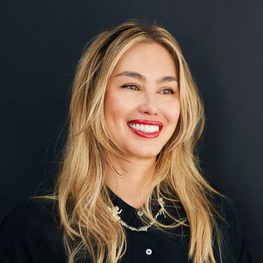 A young woman with long blonde hair, smiling with bright red lipstick, wearing a black top and a chain necklace, against a dark background.