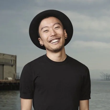 A young man with a big smile, wearing a black wide-brimmed hat, black t-shirt, earrings, and ear gauges, standing outdoors near the water with an industrial background and cloudy sky.