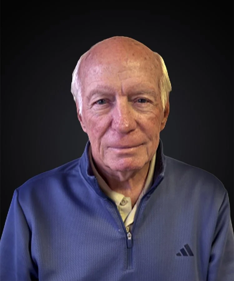 Head and shoulders portrait of an elderly man with light skin and short, light-colored hair, wearing a navy blue zip-up jacket over a light-colored collared shirt, against a dark background.
