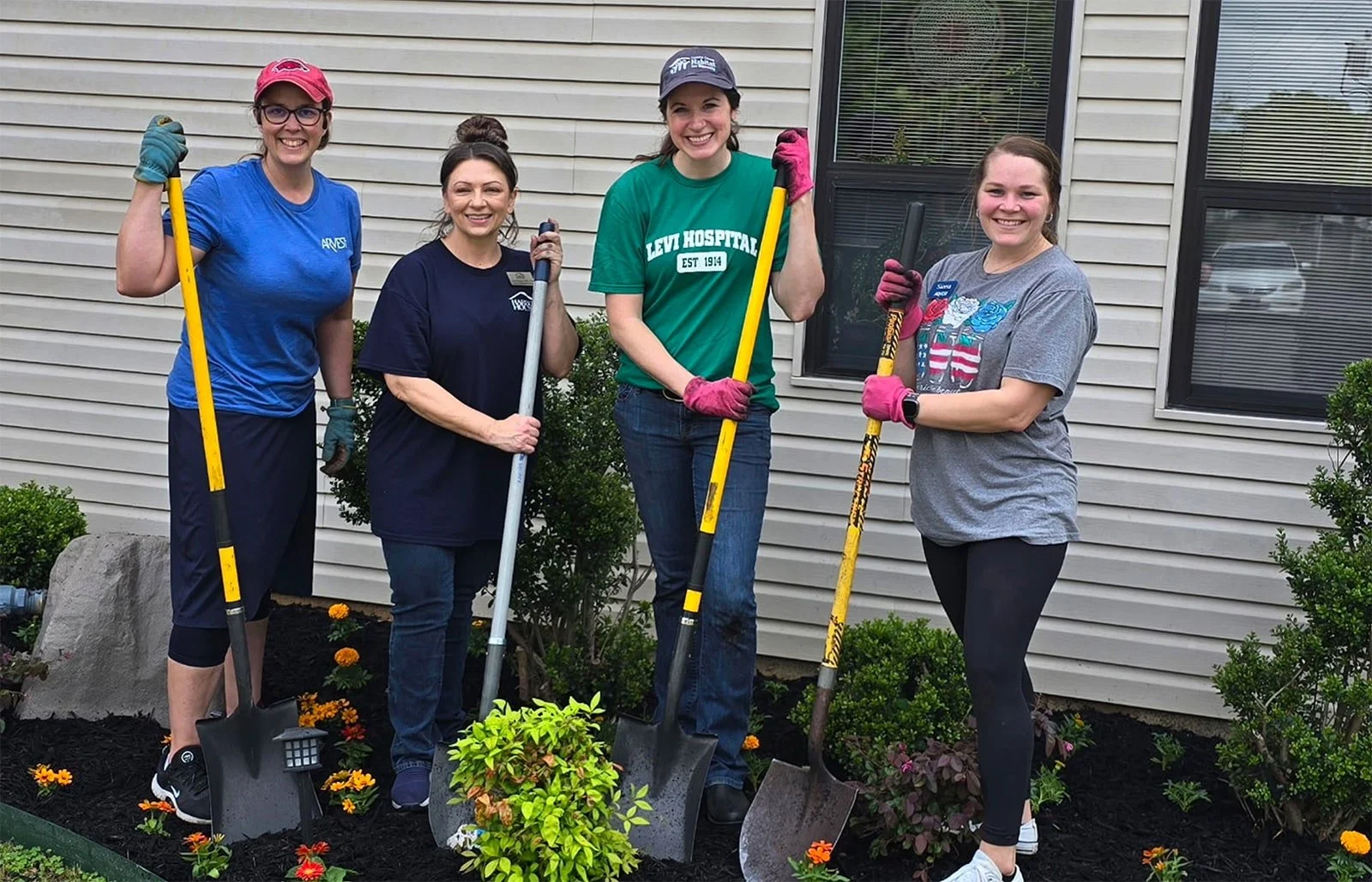 Four women standing outdoors in front of a house, holding gardening shovels, smiling, with flower bed and bushes around them.