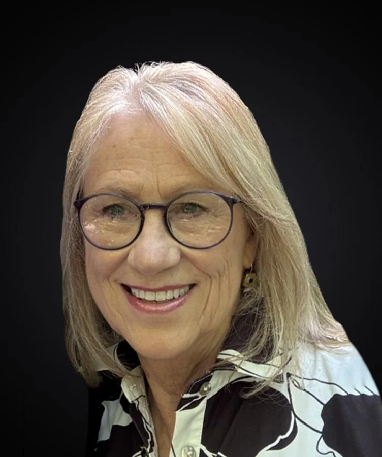 A woman with shoulder-length blonde hair and glasses, smiling against a black background.