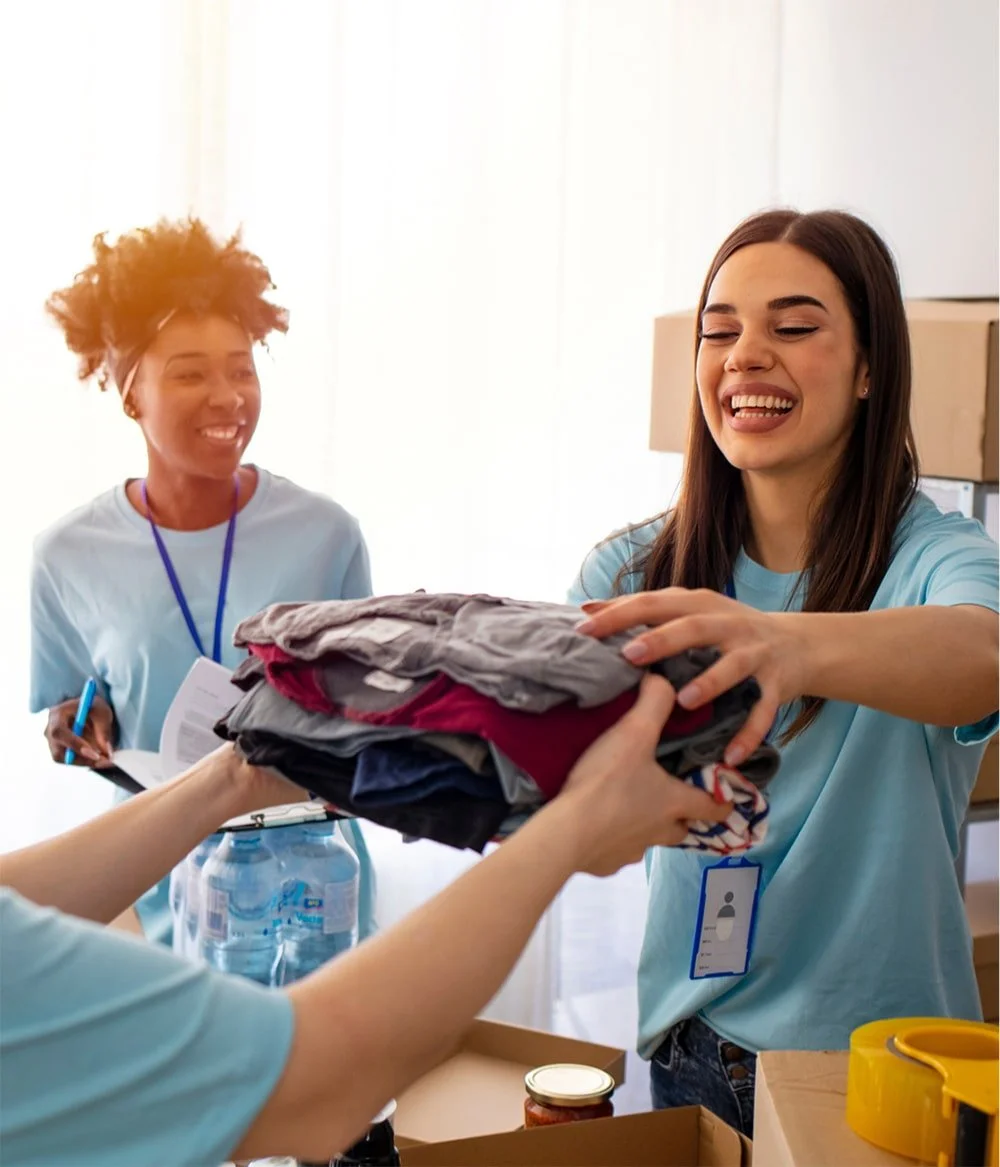 Two women in light blue shirts, smiling, are handing over a pile of folded clothes to a person whose hands are visible. The background shows cardboard boxes and supplies, suggesting a donation or volunteer setting.
