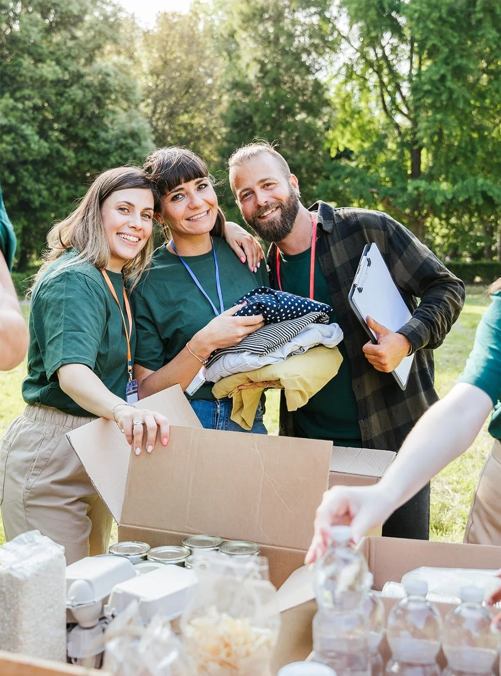 Three happy people with volunteering badges are smiling at the camera while holding folded clothes. They are outdoors in a park, with a table in front of them filled with food and supplies, participating in a community service or charity event.