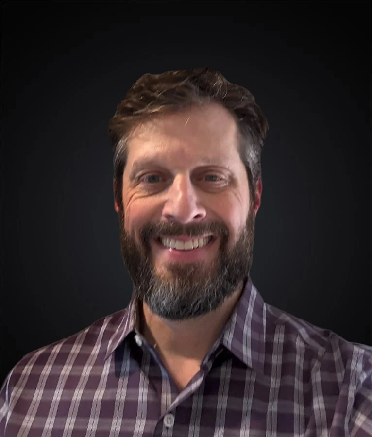 A man with brown hair and a beard smiling, wearing a purple checkered shirt, against a dark background.