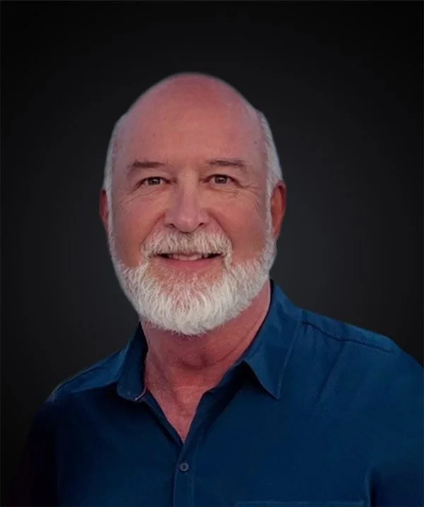 Portrait of a smiling older man with a white beard, wearing a dark blue collared shirt, against a dark background.