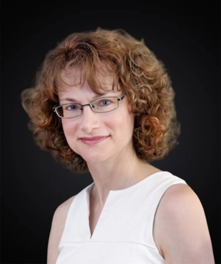 A woman with curly red hair wearing glasses and a sleeveless white top, smiling against a dark background.