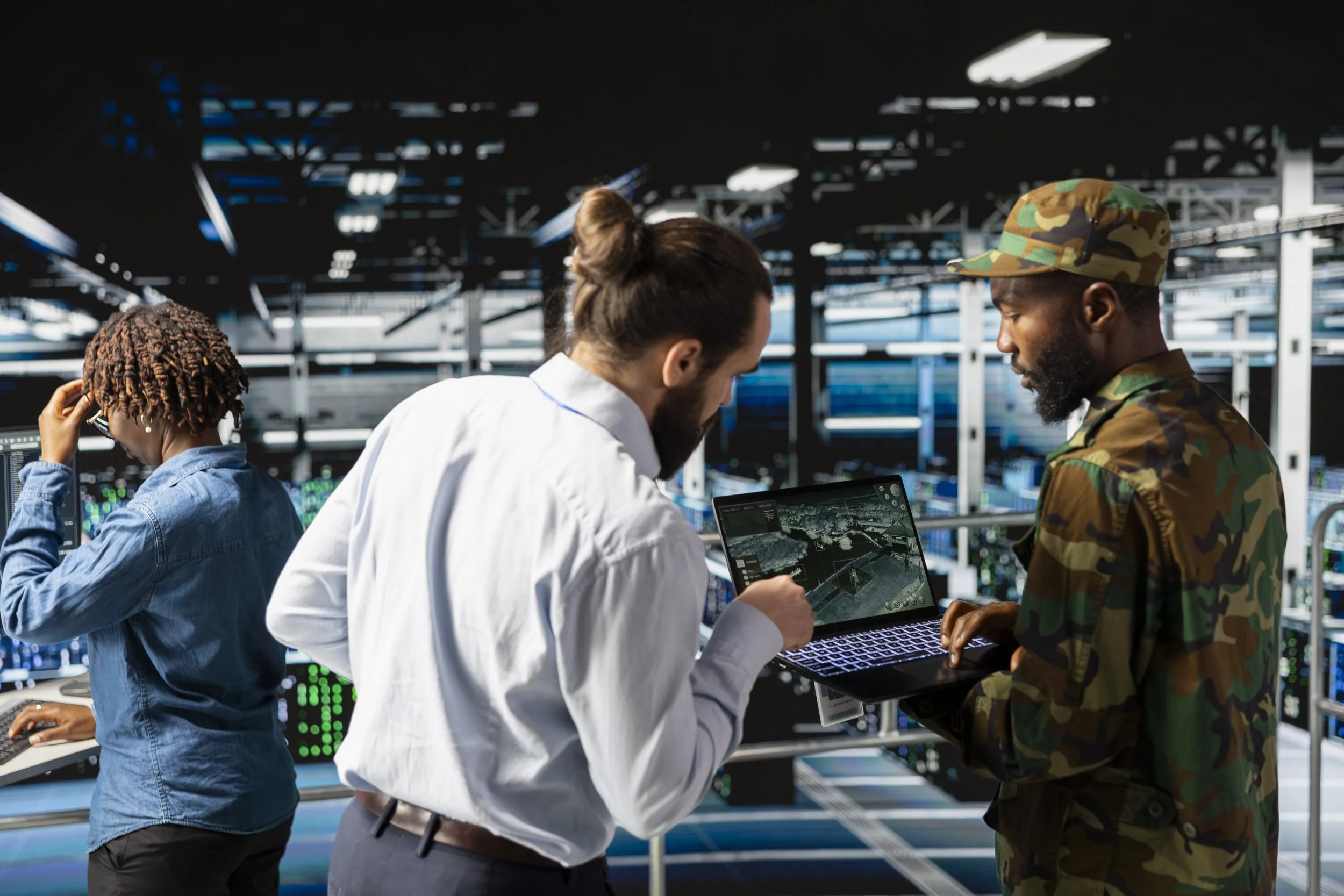 Three individuals working in a high-tech data center, with one person in casual jeans and another in a white shirt, and a third person in military camouflage, analyzing data on a laptop.