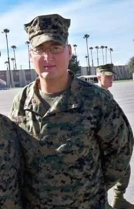 A person in military camouflage uniform and cap standing outdoors with palm trees in the background, alongside another person in similar uniform.