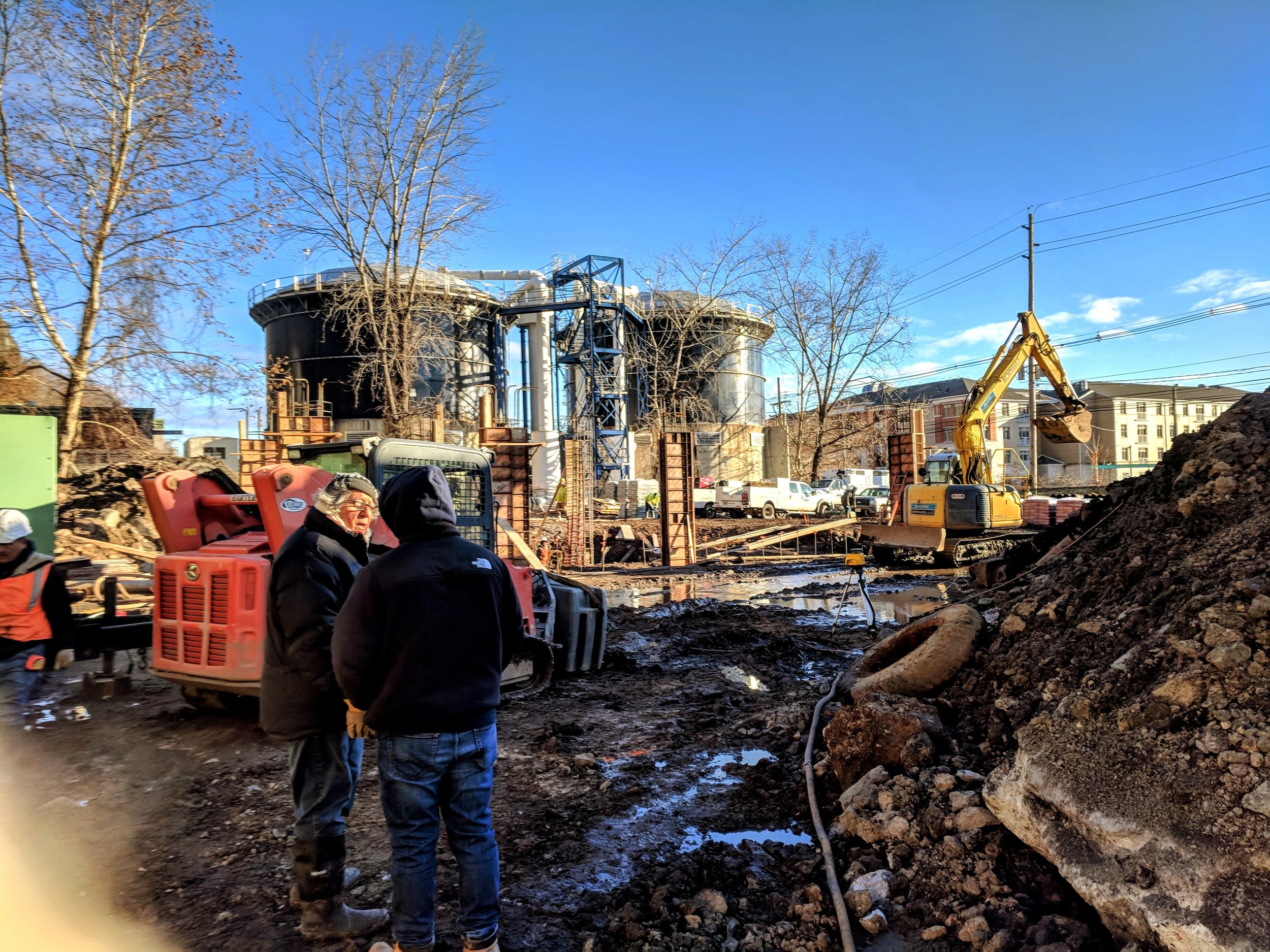 Construction site with two men talking in the foreground, construction equipment, muddy ground, and large industrial structures under a clear blue sky.