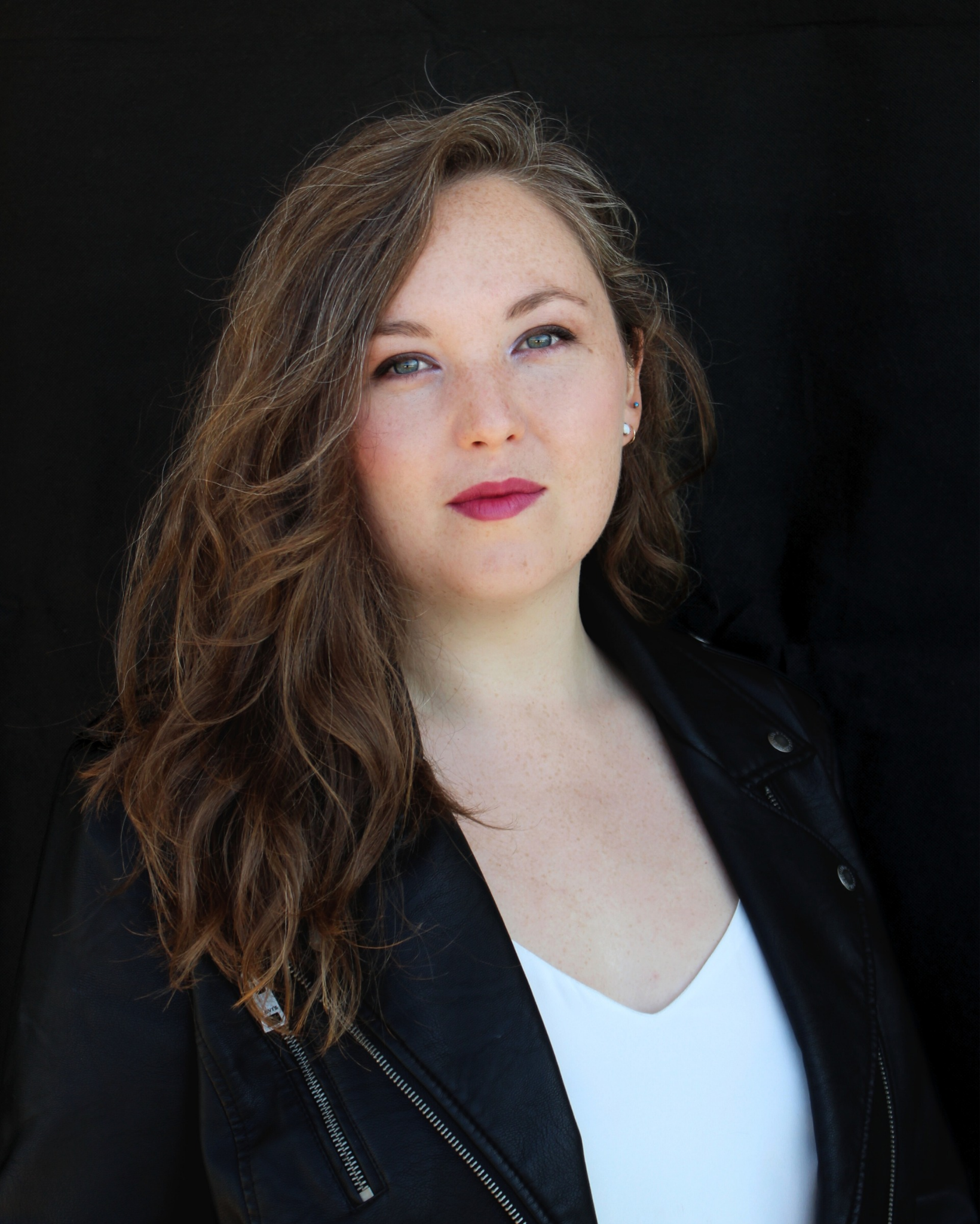 A woman with long, wavy brown hair, blue eyes, and light skin with freckles, wearing a black leather jacket over a white top, standing against a black background.