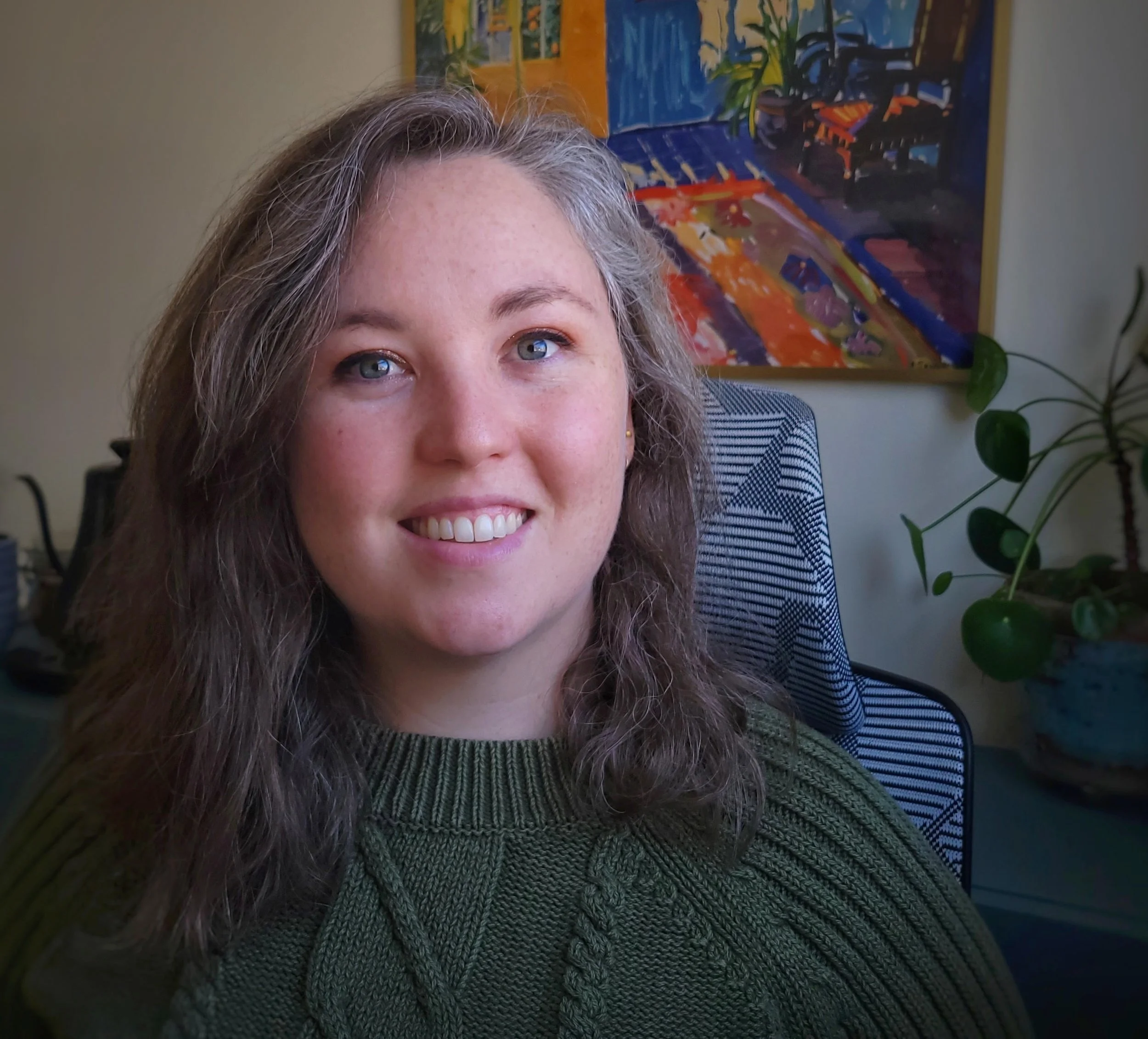 Close-up of Morgan Schreiber with long curly hair, smiling, wearing a green knit sweater, sitting in an office with colorful abstract painting in the background and a houseplant to her right.