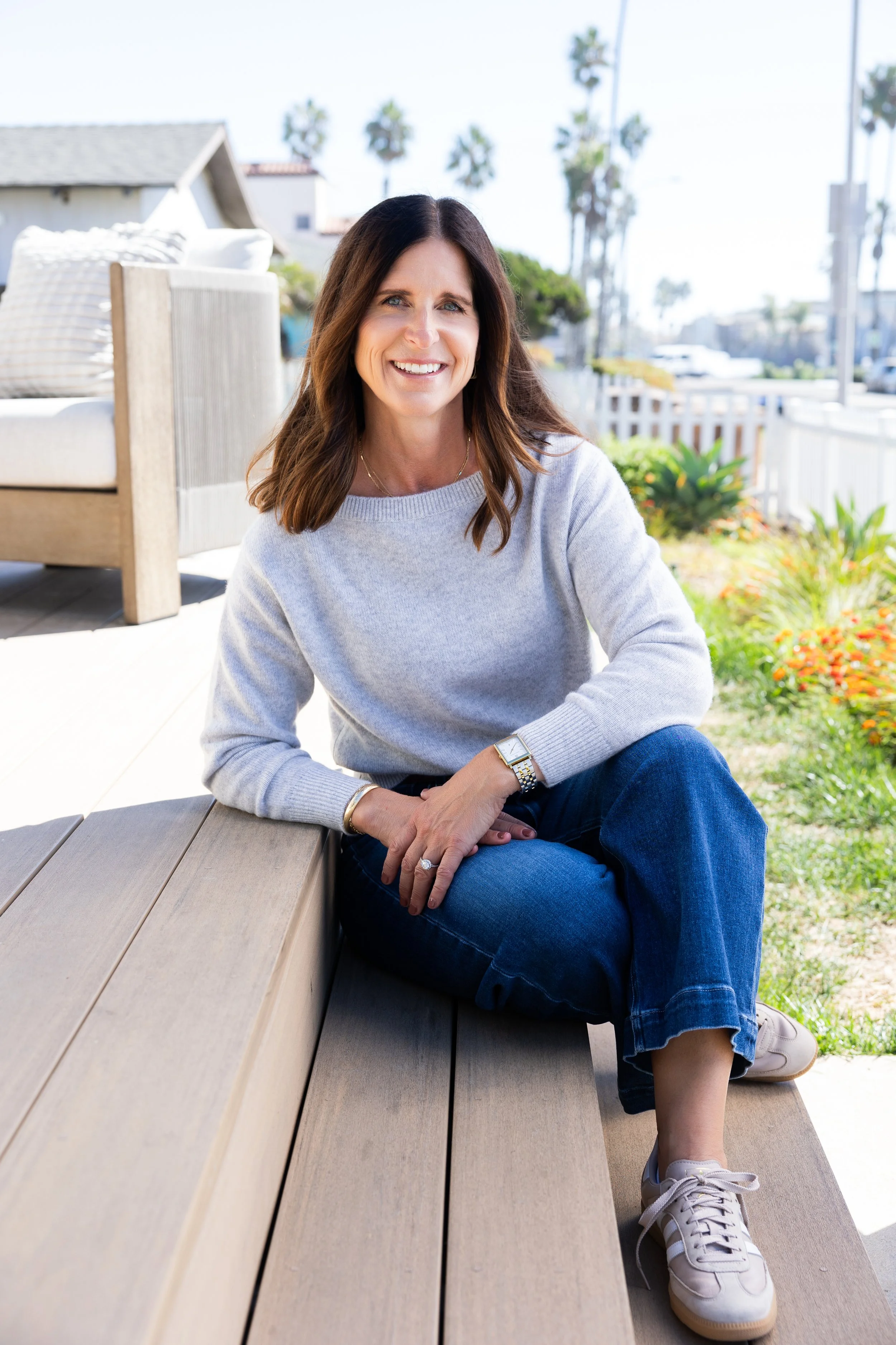 Smiling woman sitting outdoors on a bench, wearing a gray sweater, jeans, and sneakers, with palm trees and a porch in the background.