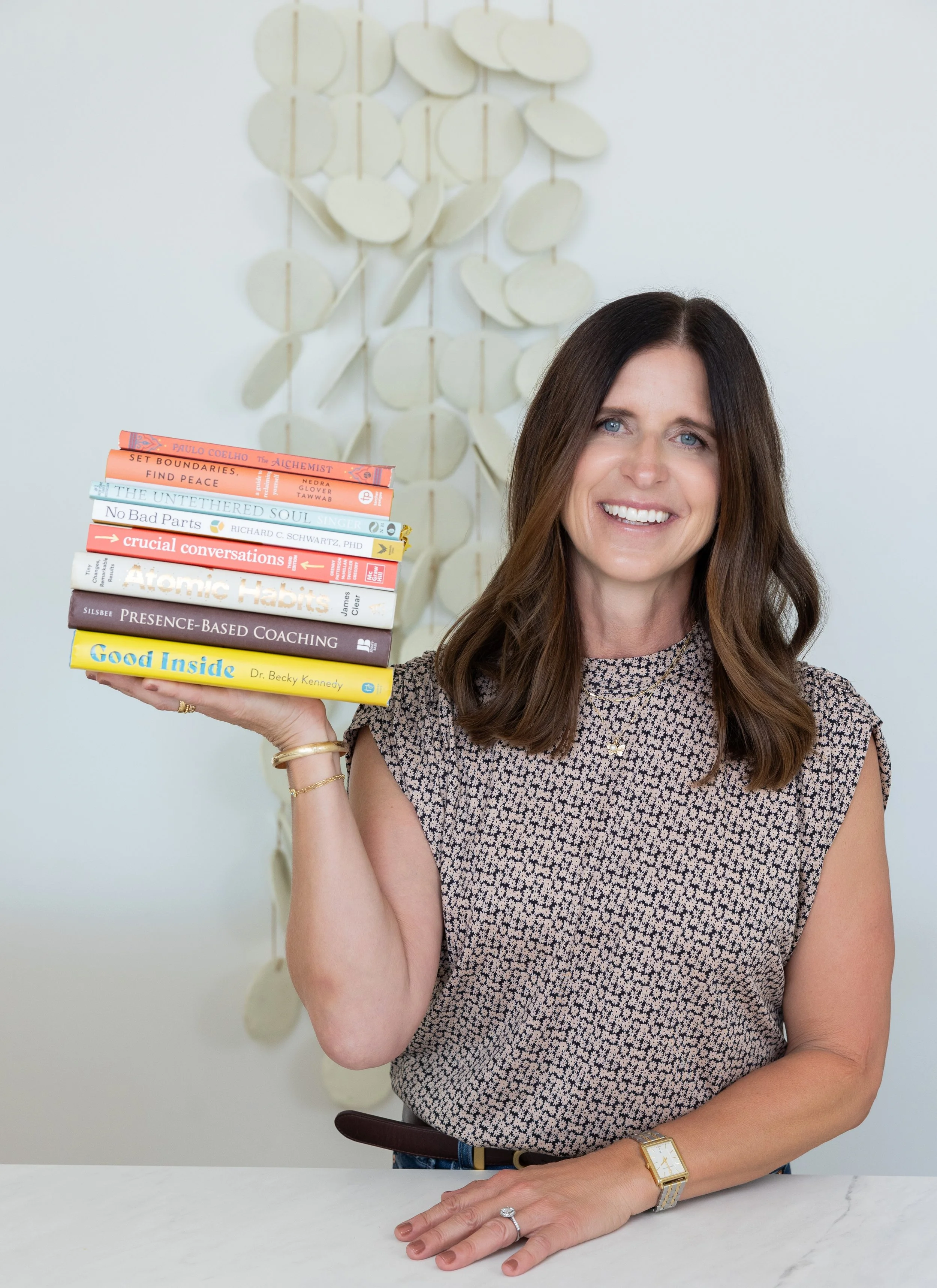 A woman smiling and holding a stack of books in a bright room with white walls and a decorative wall hanging behind her.