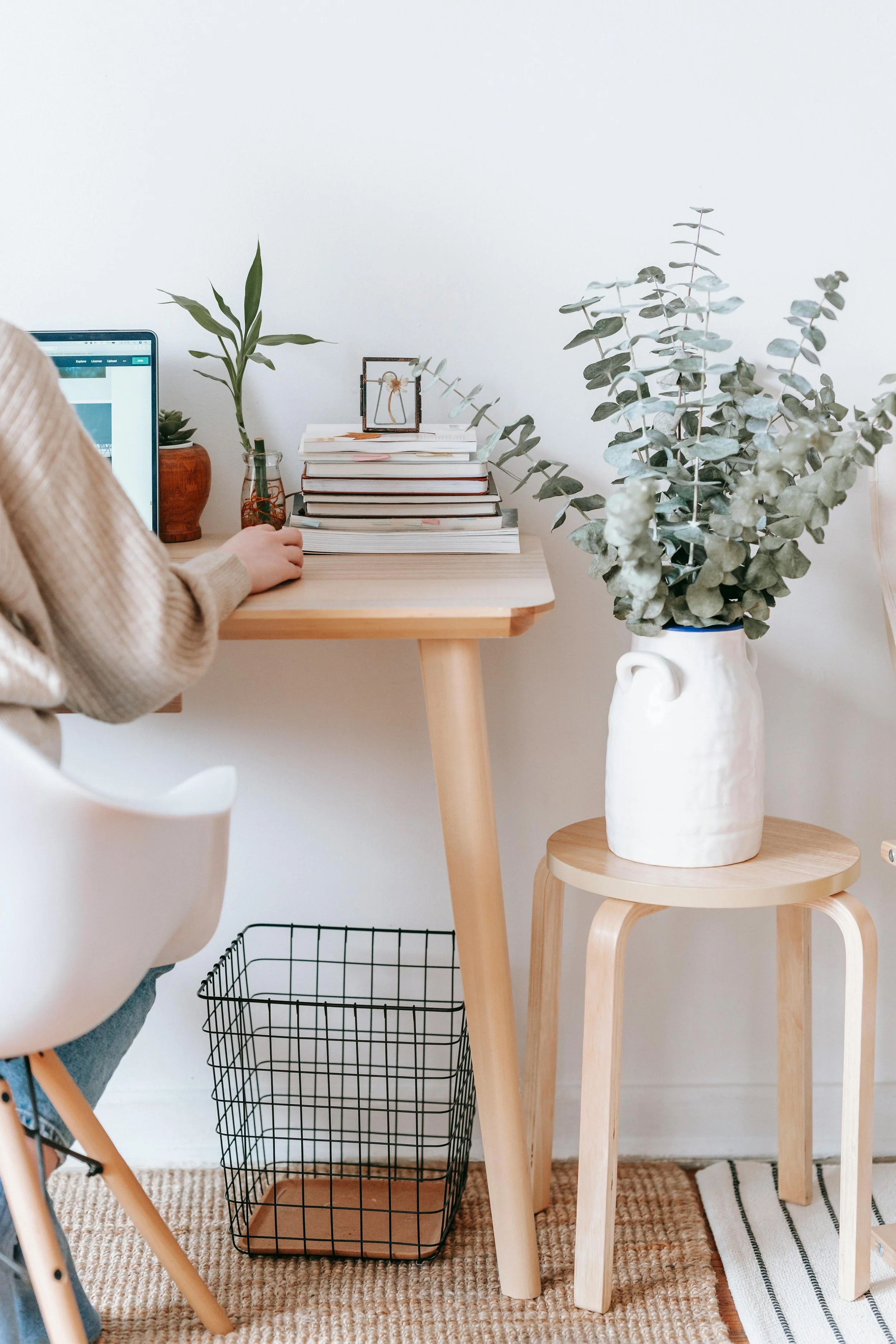 A workspace with a light wood desk, a white chair, a laptop, a small potted plant, a glass of water, potted plants, a stack of books, a framed photo, a large vase with eucalyptus, a small wooden stool, a wire basket, a striped rug, and a white wall.