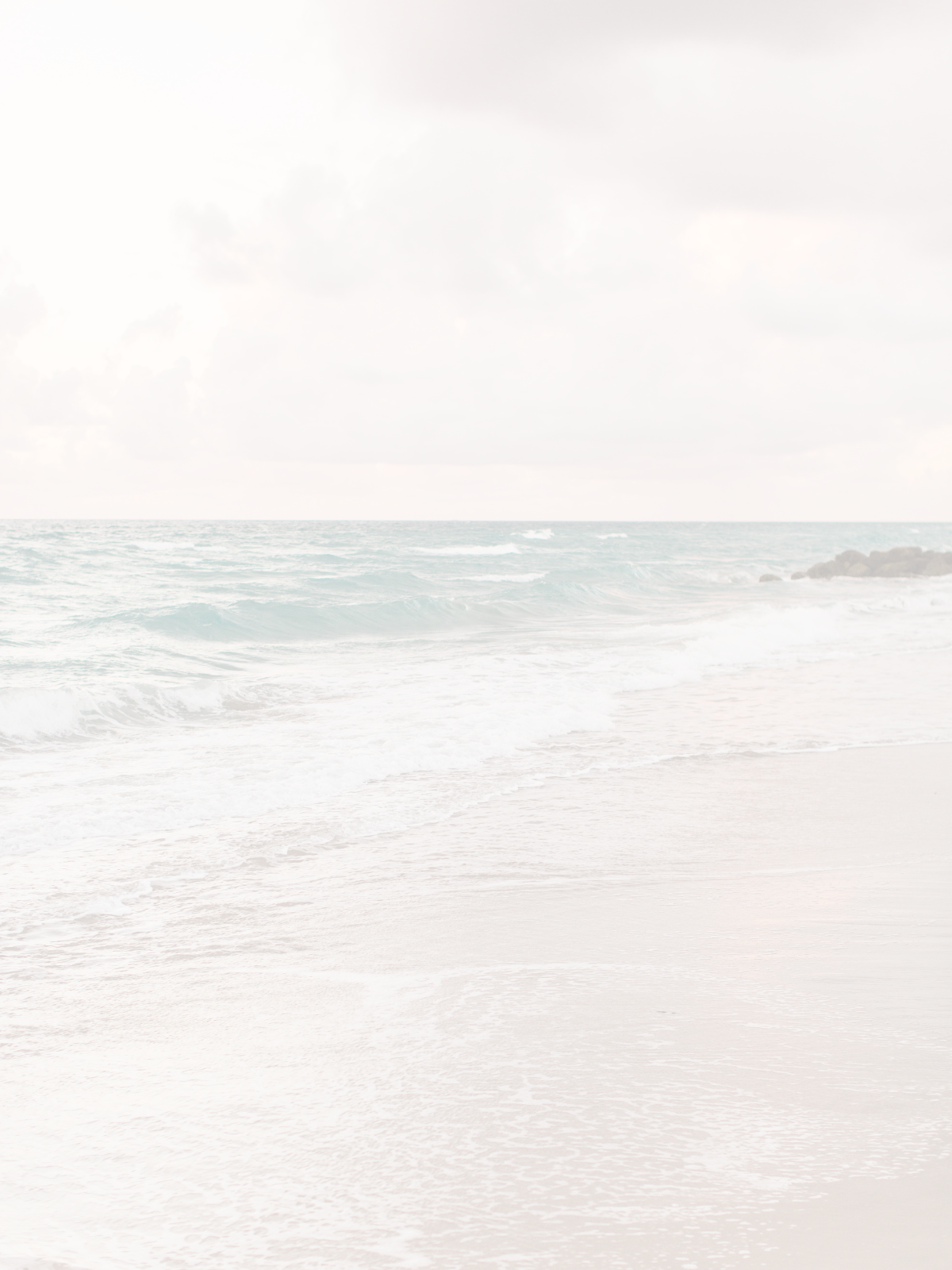 A serene beach scene with gentle waves on the shoreline, light-colored sand, and a cloudy sky.