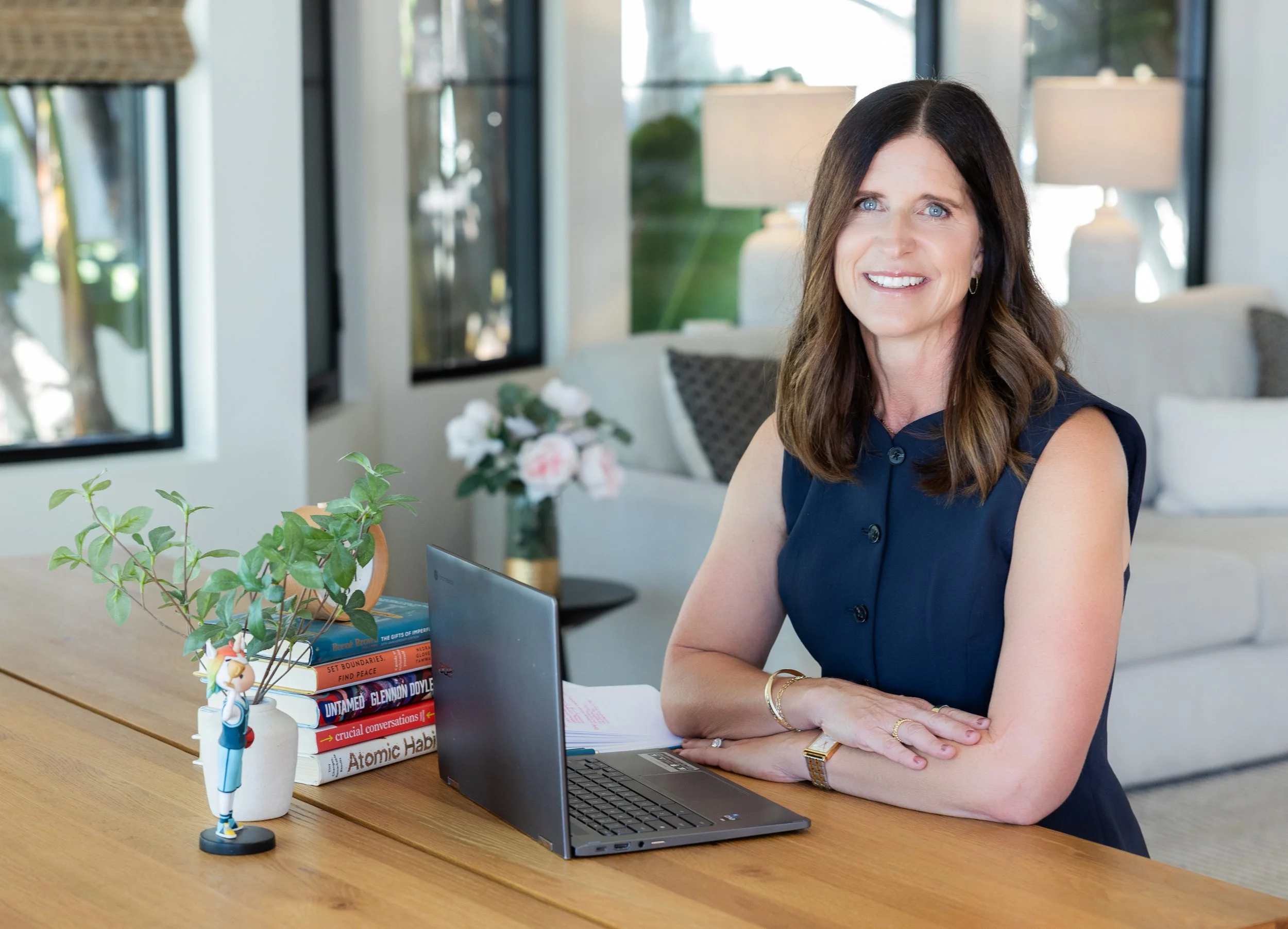 A woman with brown hair and a navy blue sleeveless dress sitting at a wooden desk, smiling. On the desk are a small potted plant, a stack of books, a laptop, and a small figurine. The background shows a living room with large windows, lamps, and a couch with pillows.