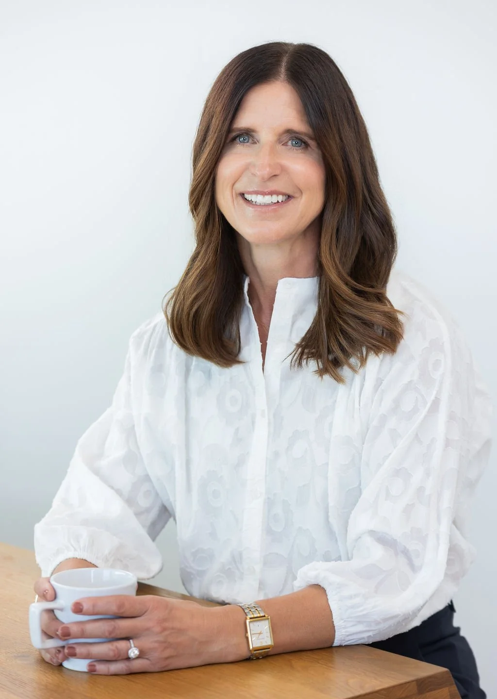A woman with brown hair and blue eyes smiling, seated at a wooden table, holding a white mug, wearing a white blouse and a gold watch.