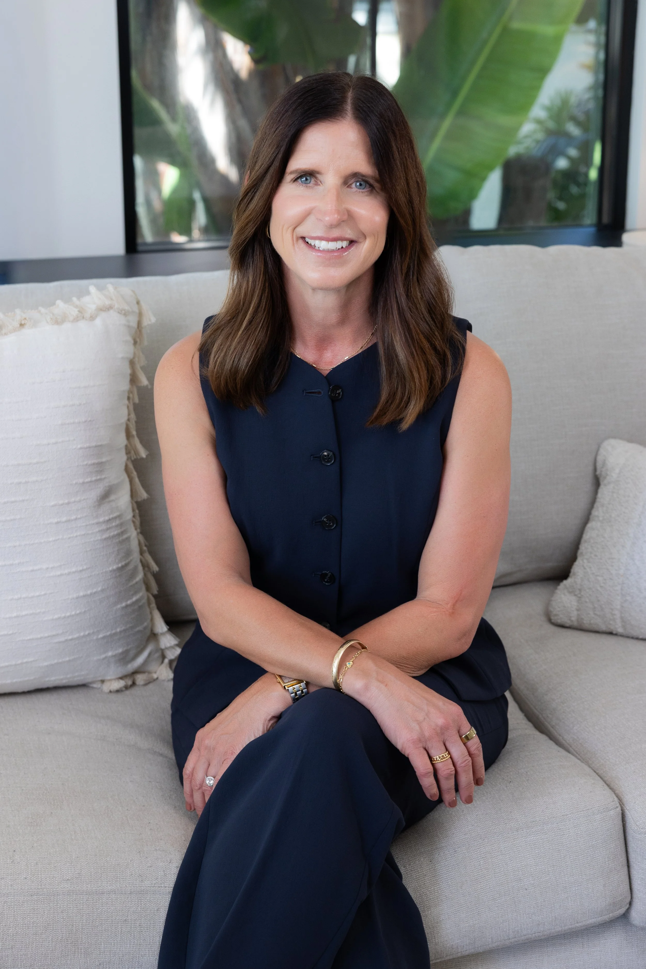 A woman with shoulder-length brown hair and blue eyes, smiling and sitting on a light-colored sofa with textured pillows, in front of a window with a view of large green leaves outside.