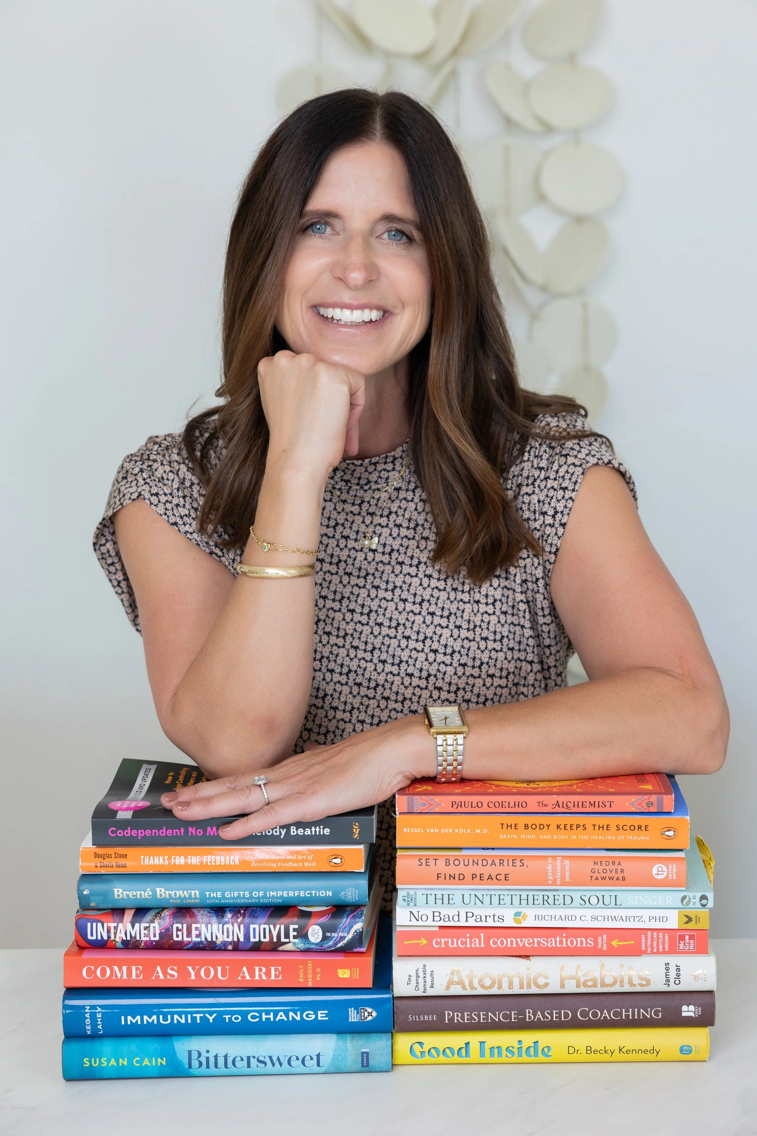 A woman with long brown hair and a patterned blouse is smiling, resting her chin on her hand, seated behind a stack of various books.