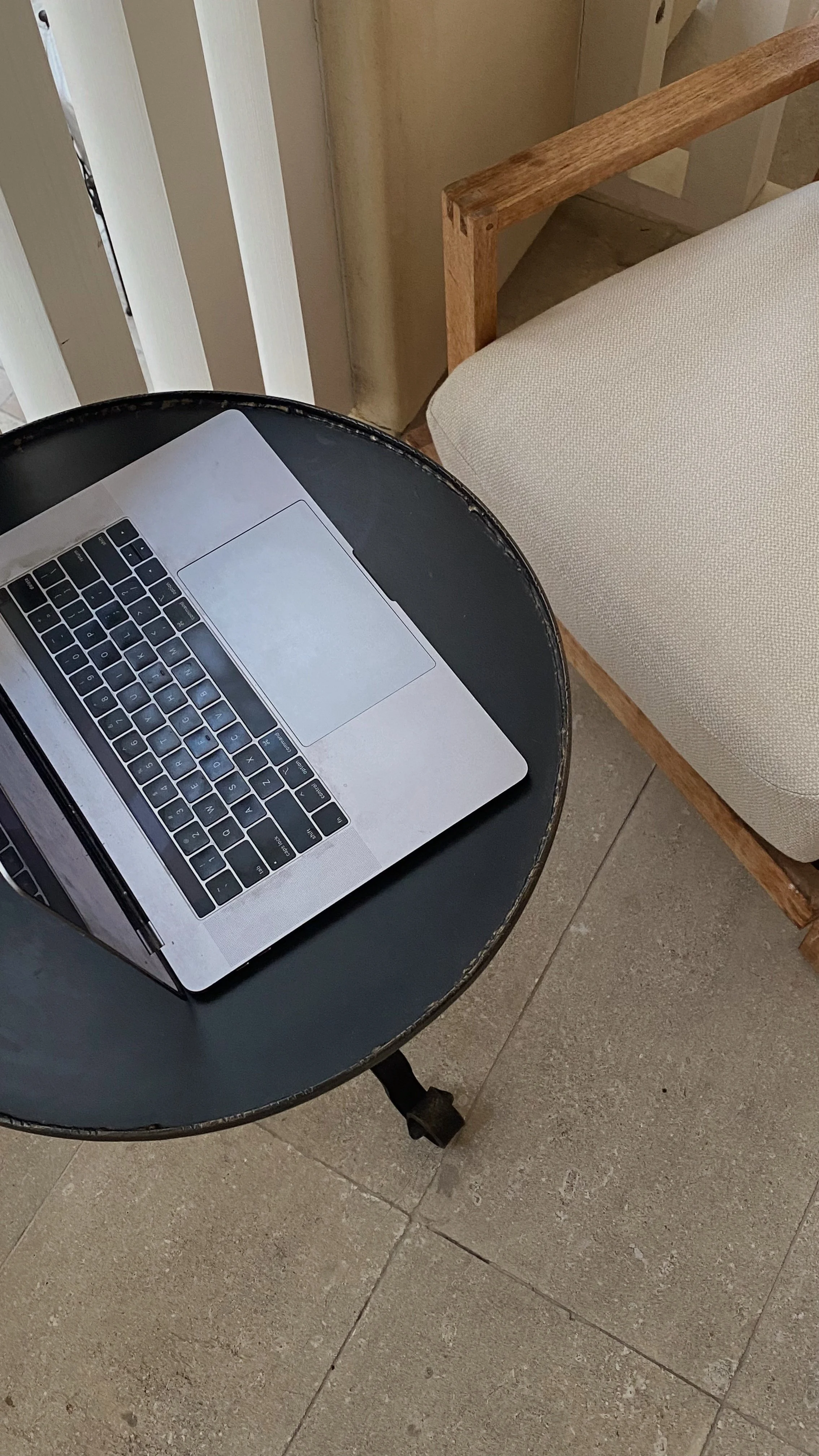 A laptop on a black round table near a cream-colored cushioned chair.