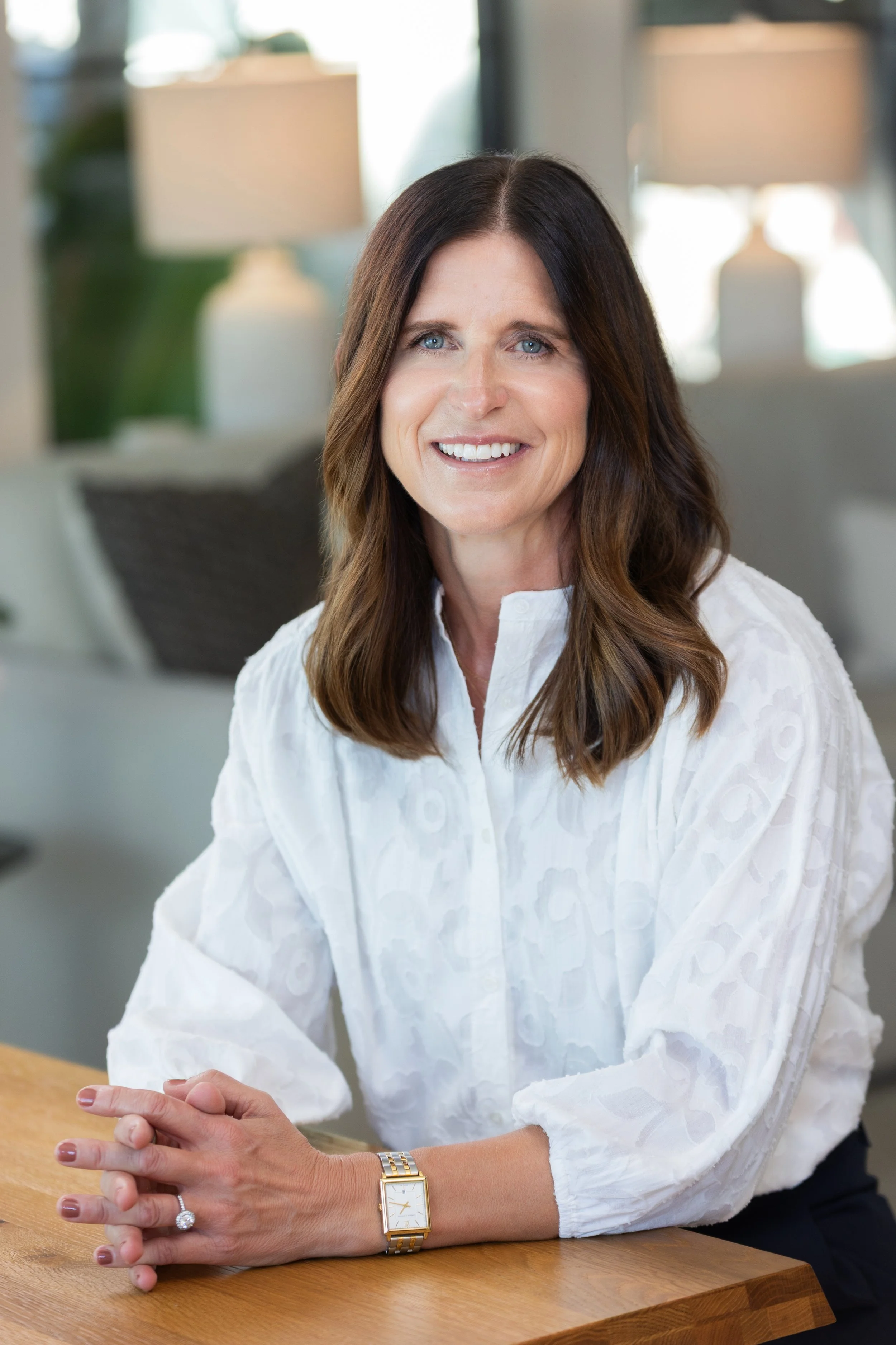 A woman with dark brown hair, blue eyes, and fair skin, smiling, sitting at a wooden table in a well-lit room, wearing a white blouse, a gold wristwatch, and a ring on her left hand.