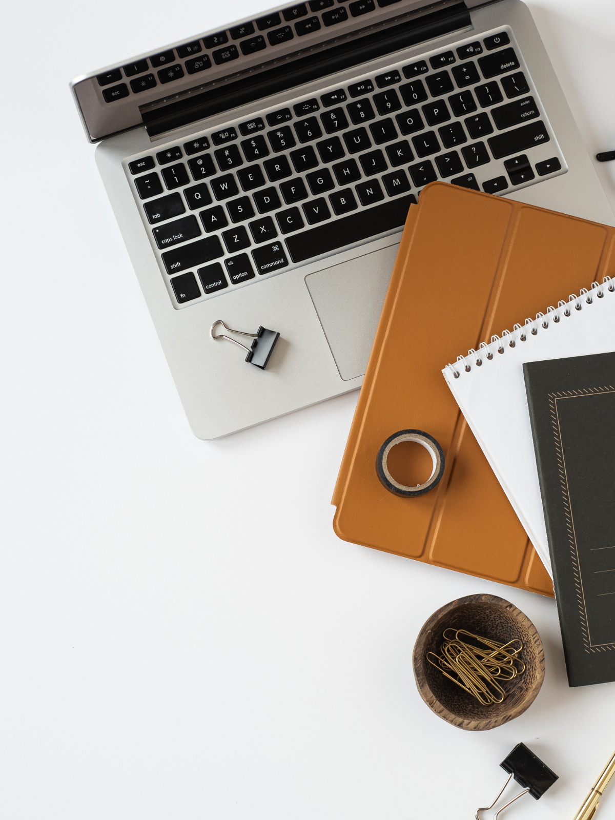 Overhead view of a workspace with a partially open laptop, a brown folder, a black notebook, notebooks, a roll of black tape, gold paper clips in a small bowl, and binder clips on a white surface.