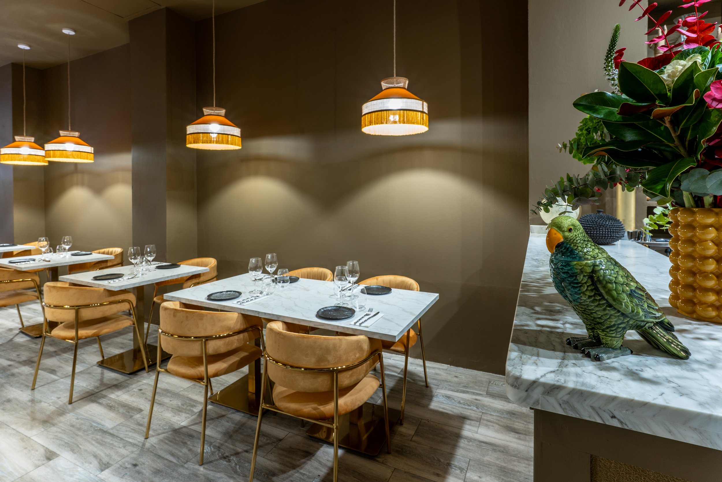 Sala principal del restaurante De Contrabando, con mesas de mármol, sillas de terciopelo mostaza, lámparas colgantes y flecos. En la barra un jarrón con un ramo y un loro decorativo.