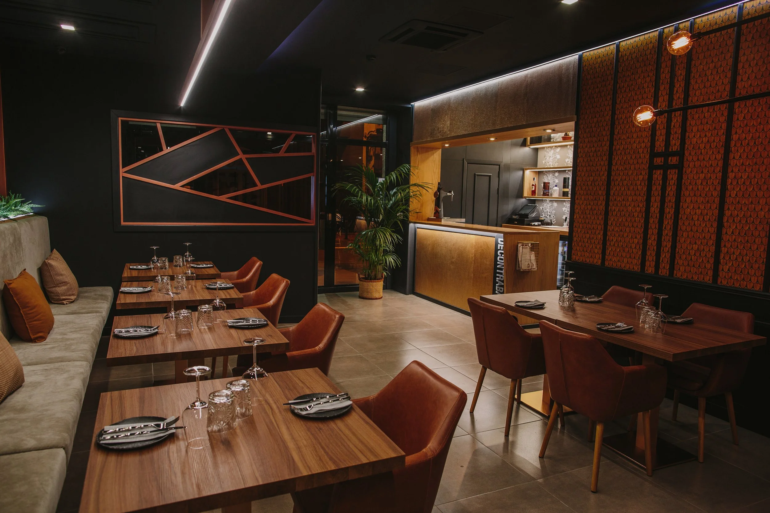Interiores de un restaurante moderno con mesas de madera, sillas de cuero marrón y decoración elegante en las paredes, con iluminación cálida y una barra de atención al fondo.