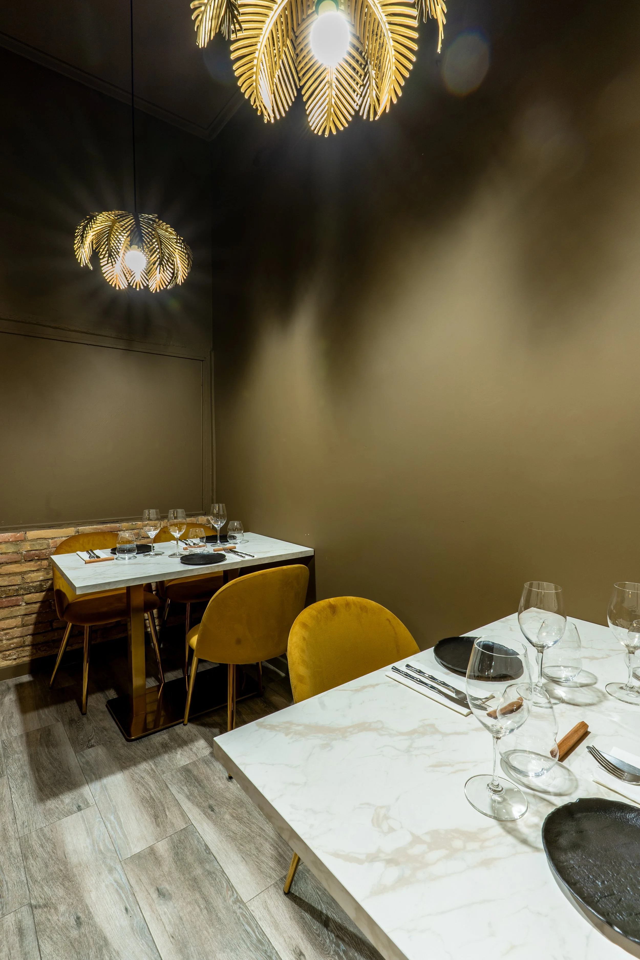 Pequeño reservado del restaurante De Contrabando, con 2 mesas de mármol blanco y sillas color mostaza. Lámparas colgantes de diseño en forma de hojas doradas.