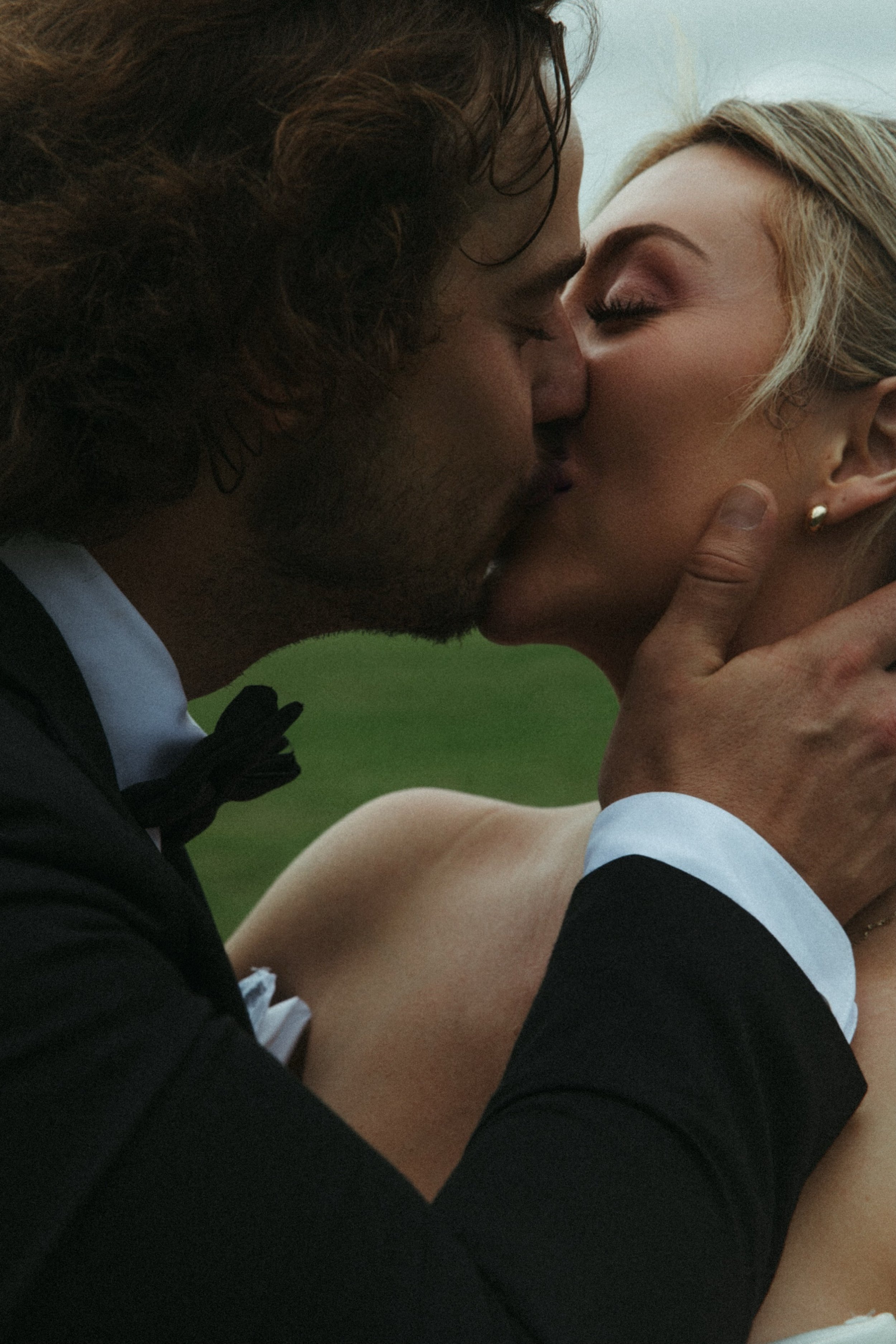 A couple dressed in wedding attire sharing a kiss outdoors, with a green background.