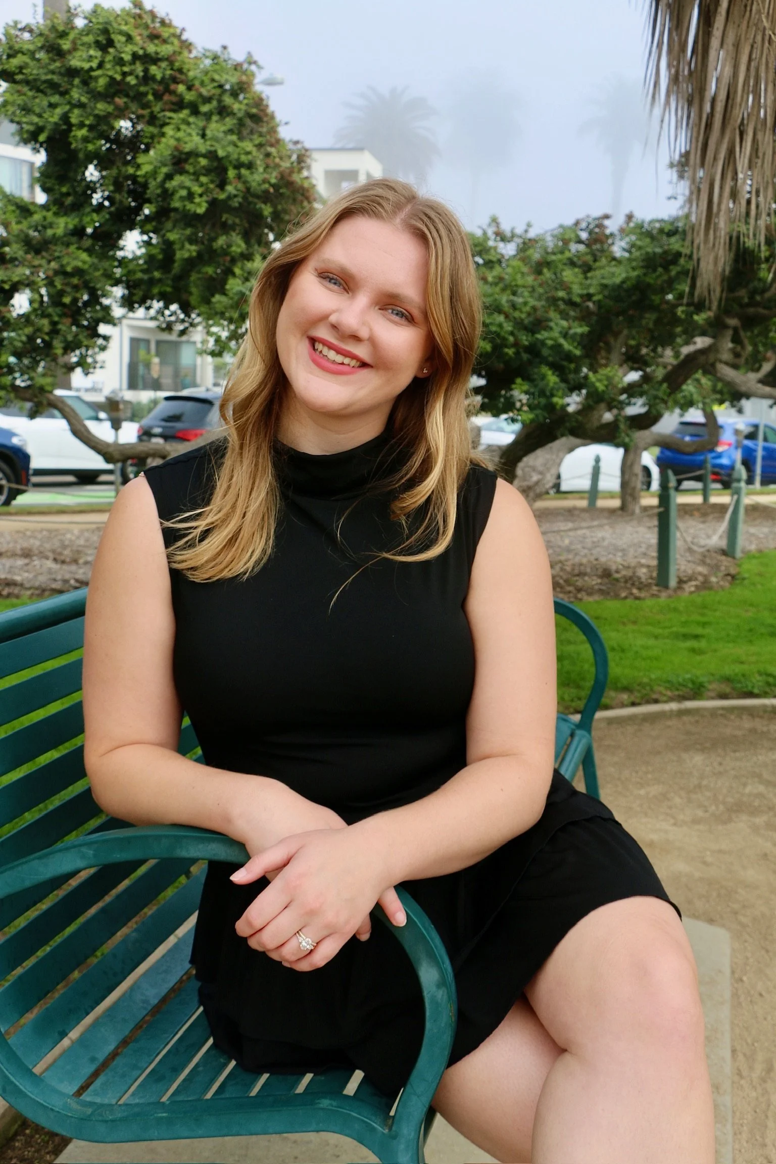 A young woman with blonde hair and fair skin, smiling, sitting on a green park bench outdoors on a cloudy day.