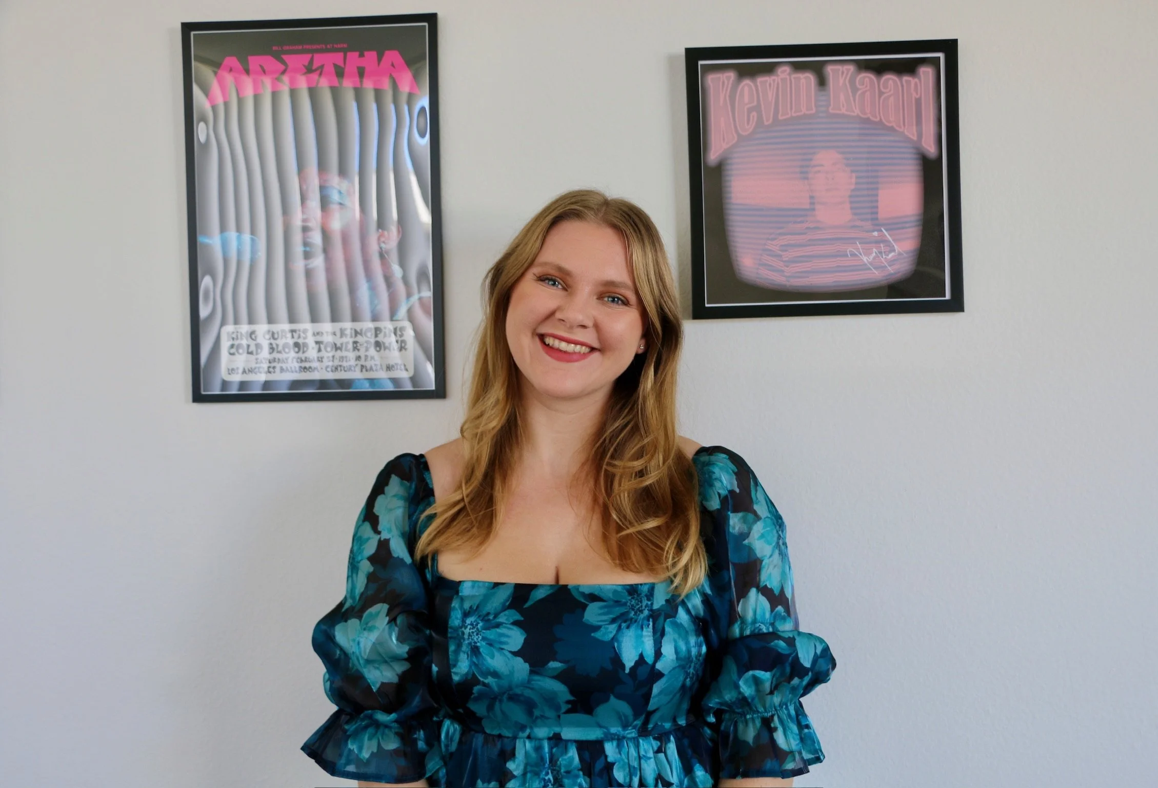 A smiling woman with blonde hair wearing a blue floral dress, standing in front of a white wall with two framed posters. The poster on the left features a person with a pink banner reading 'ARETHA' and the poster on the right shows a person with a pink neon sign reading 'Kevin Kaarl'.