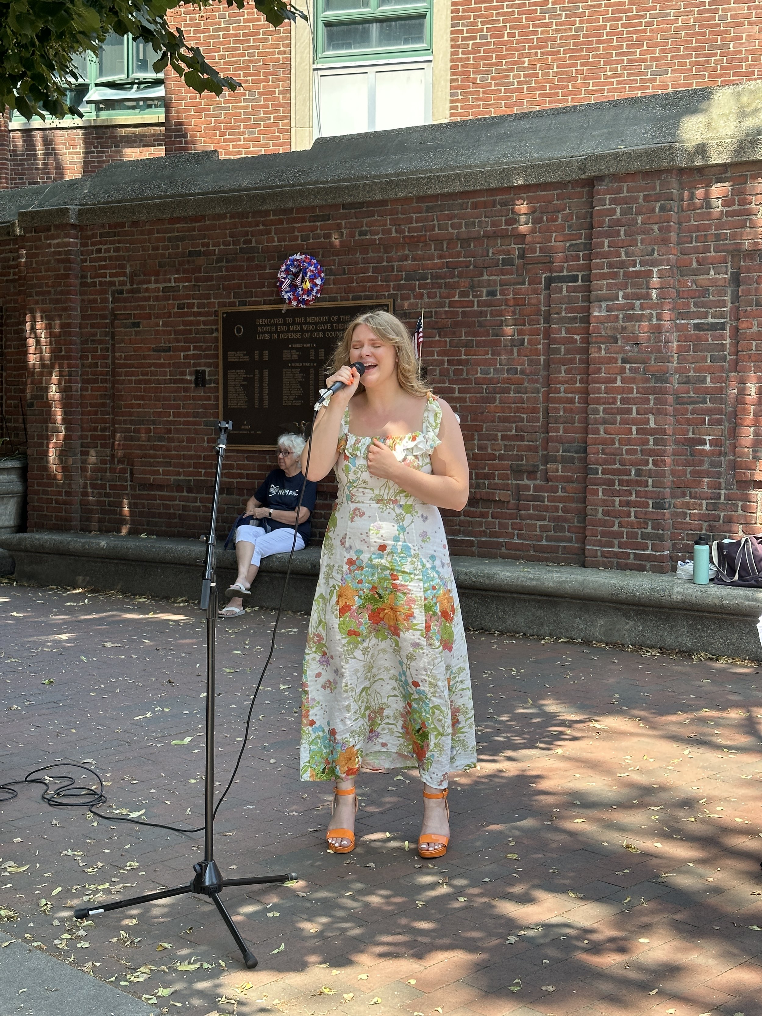 A woman singing into a microphone outdoors in front of a brick wall, with a person sitting on a bench in the background and some bags and water bottles on the ground.