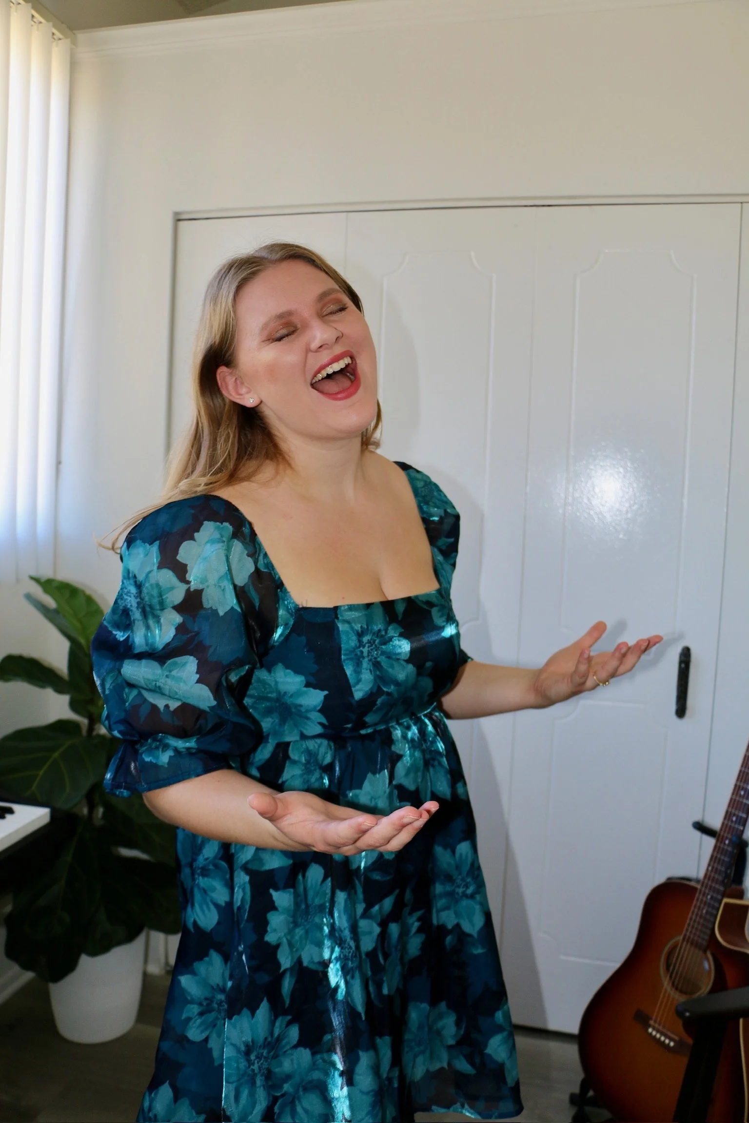 A woman in a blue floral dress laughing with her eyes closed, standing indoors next to a white door and potted plant, with a guitar and a keyboard nearby.